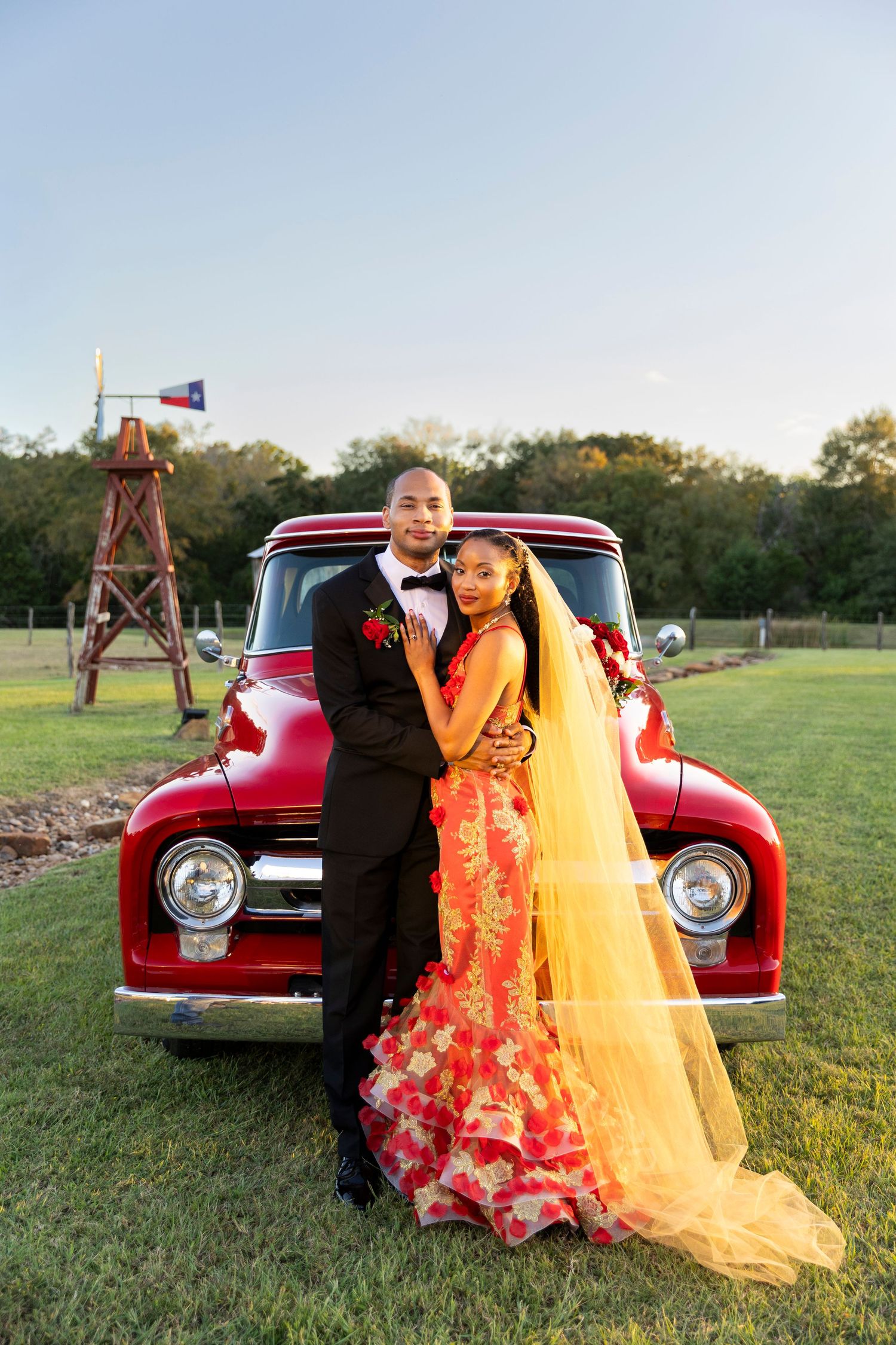 Couple in formal attire posing in front of a classic red truck at golden hour on a rural Texas property.