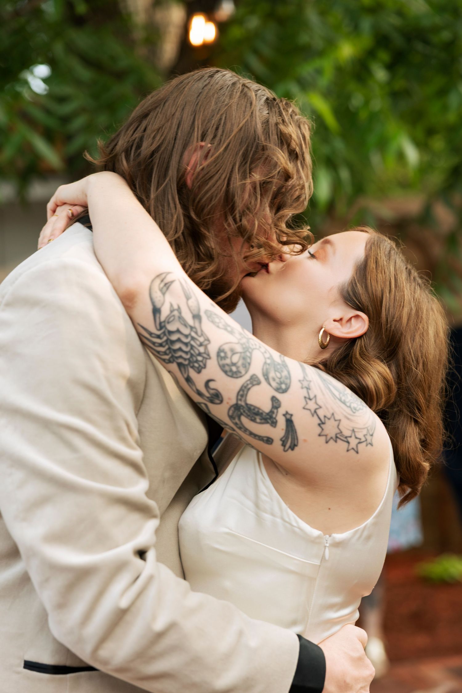 Tattooed bride and groom share a romantic first kiss outdoors at their intimate wedding ceremony.