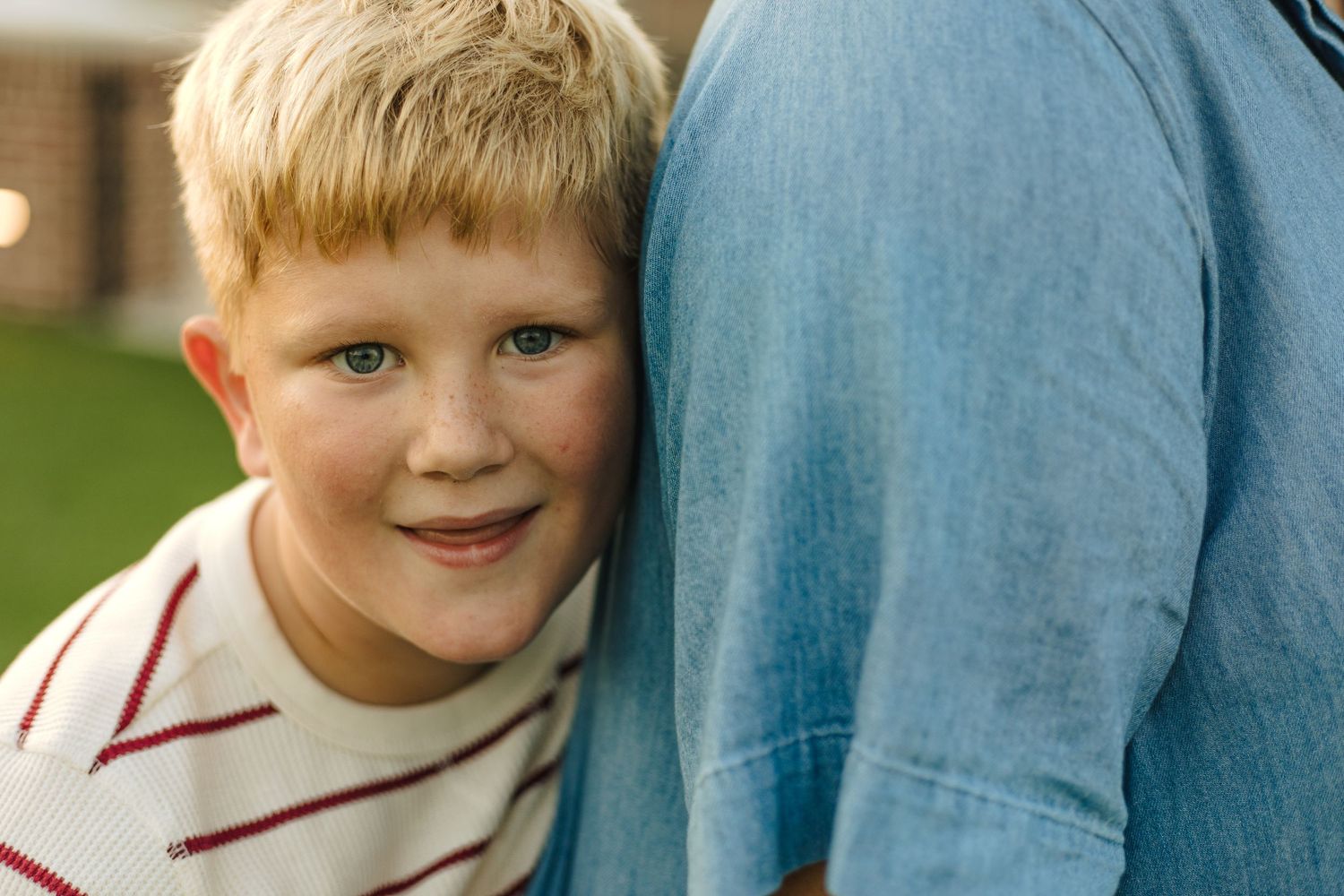 Smiling blonde boy with green eyes leaning against adult in blue jeans outdoors.