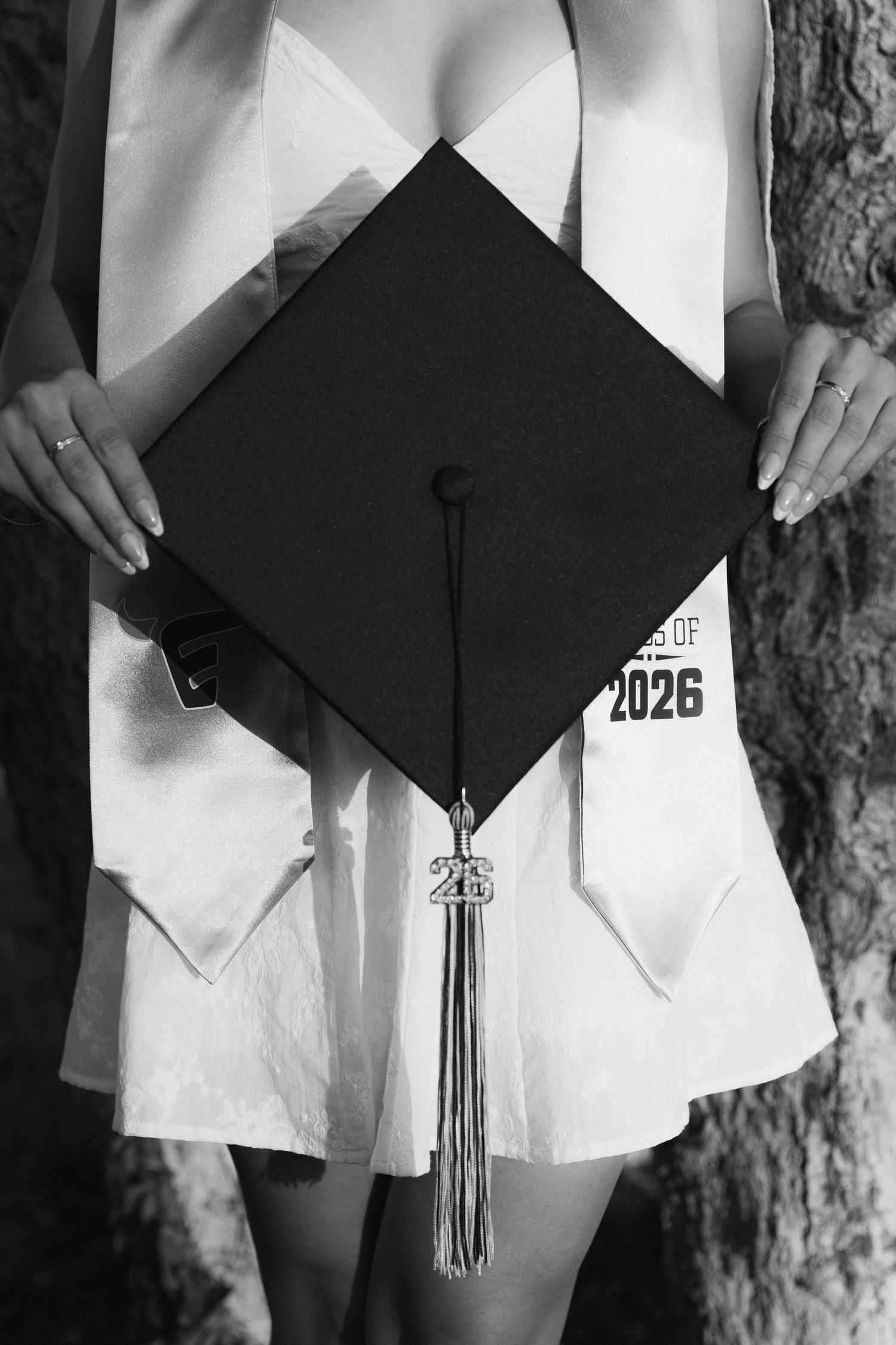 Graduate holds black mortarboard cap with Class of 2026 tassel while wearing white graduation gown.