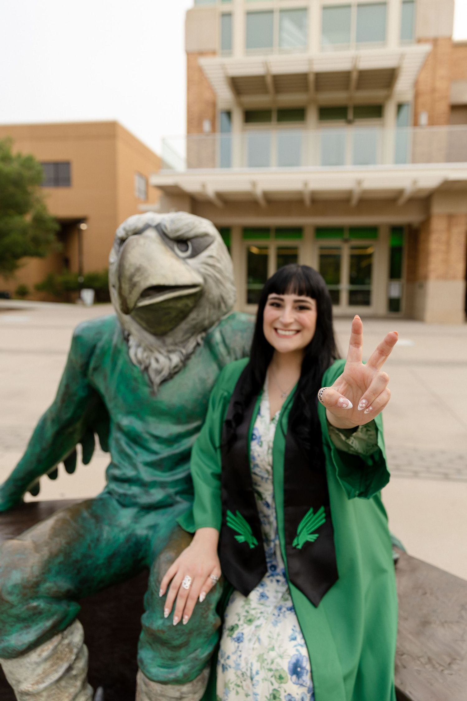 Graduate in green gown poses with eagle mascot statue, flashing peace sign at university campus.