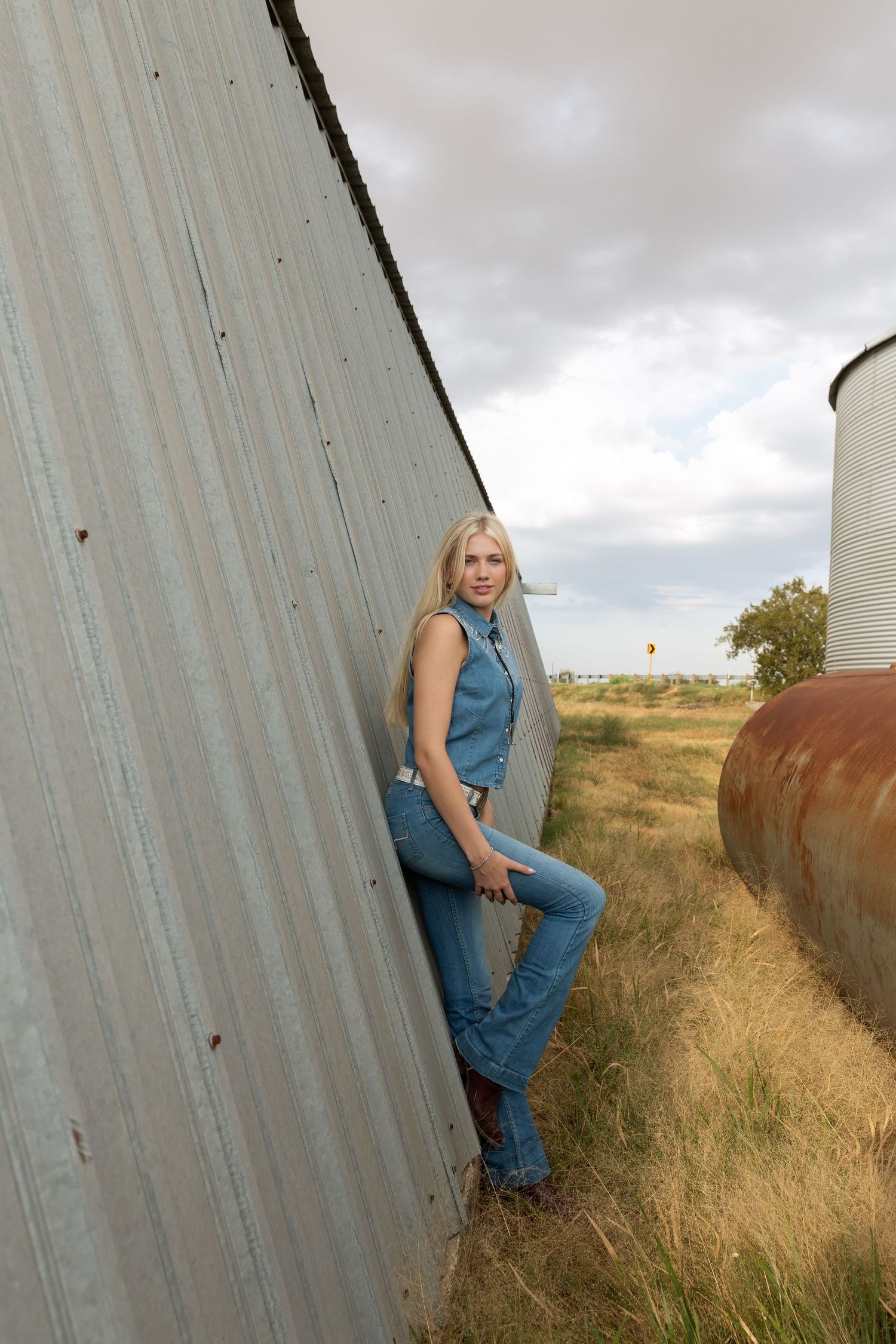 Blonde woman in denim jumpsuit leaning against rustic metal barn wall in rural farm setting with silos.