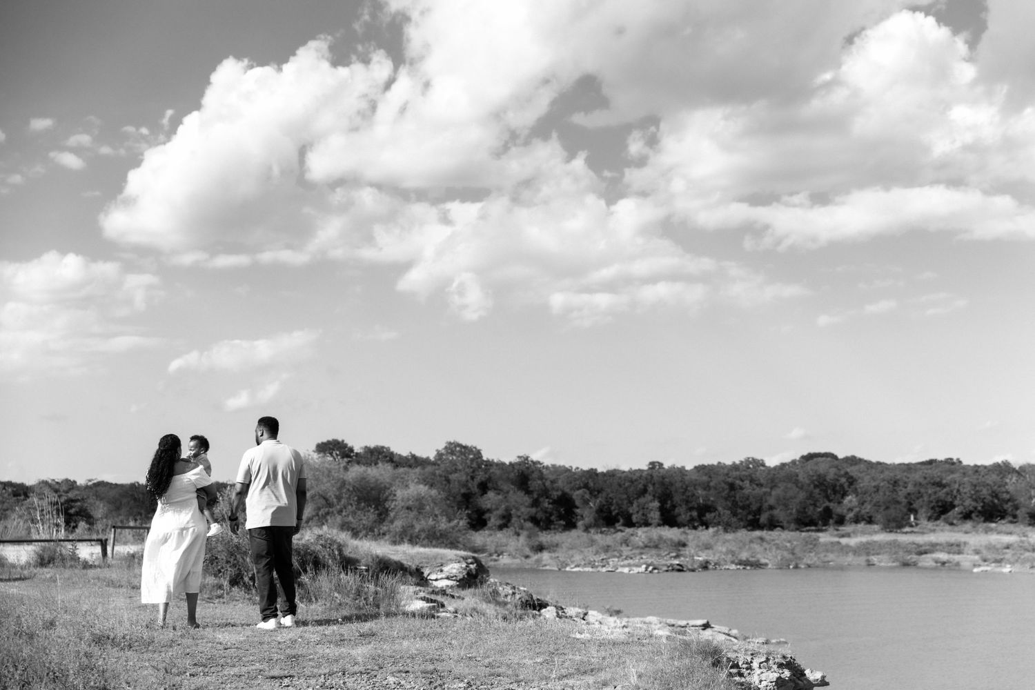 Black and white photo of couple holding hands while standing by a scenic riverside under cloudy sky.