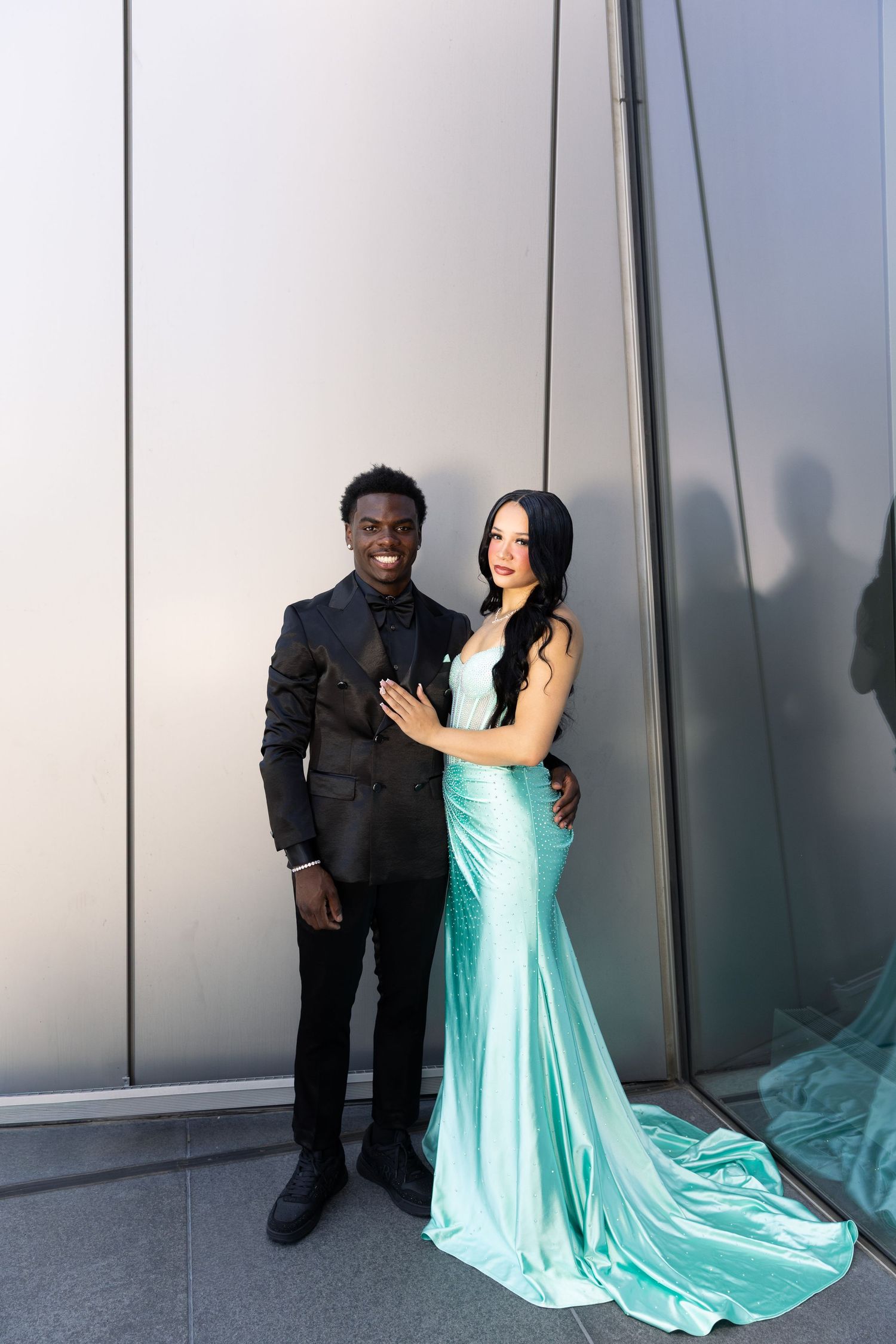 Couple posing formally in prom attire, man in black suit and woman in teal mermaid gown, against silver wall.