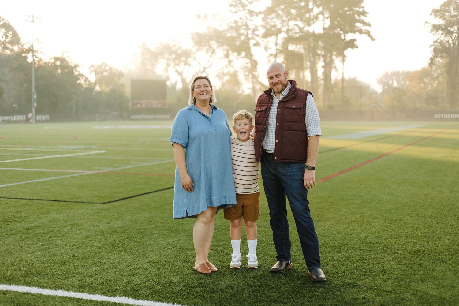 Family of three smiling together on green sports field surrounded by glowing sunset light.