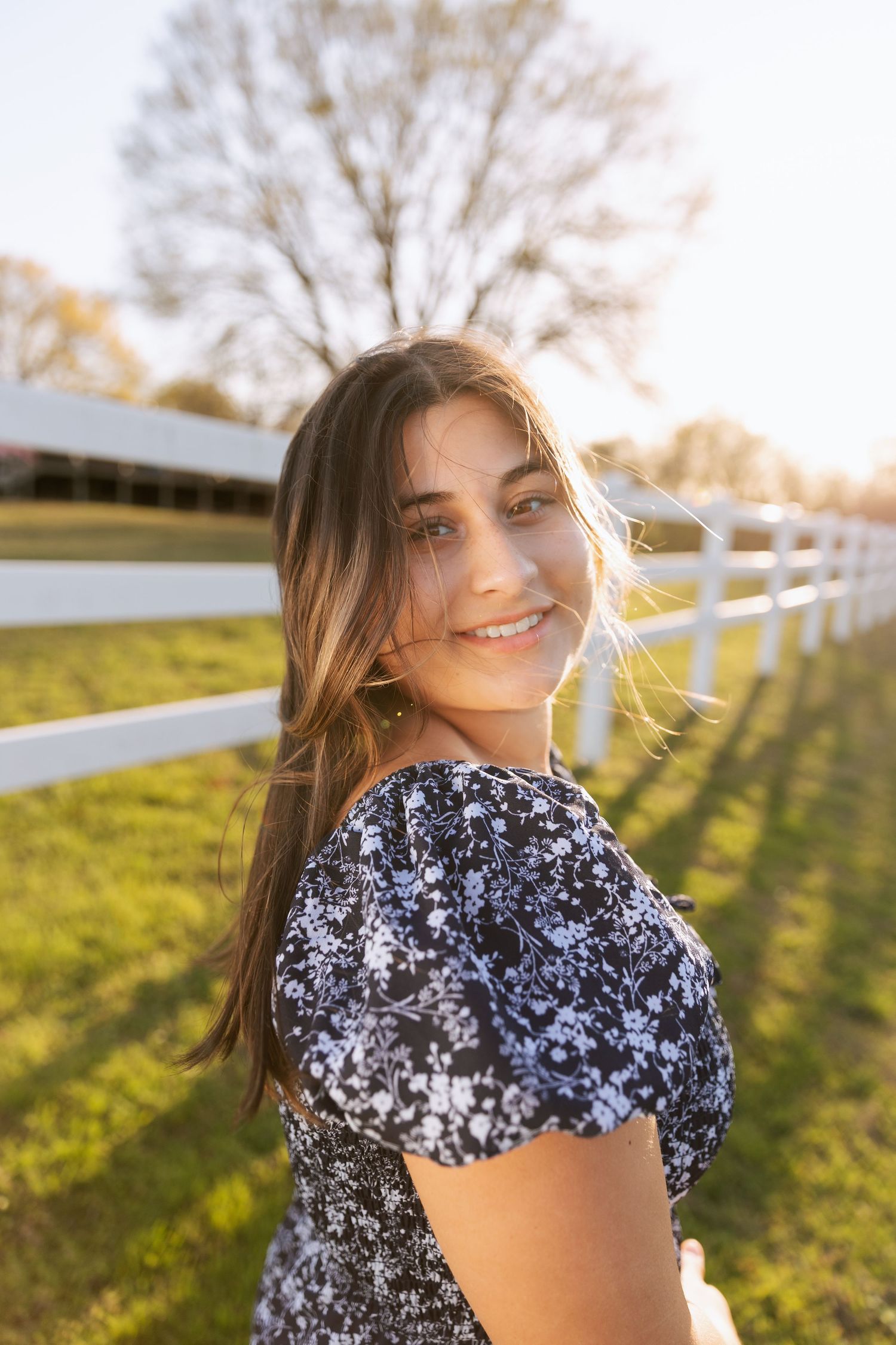 Smiling teenage girl in navy floral dress poses by white fence during golden hour senior portrait.
