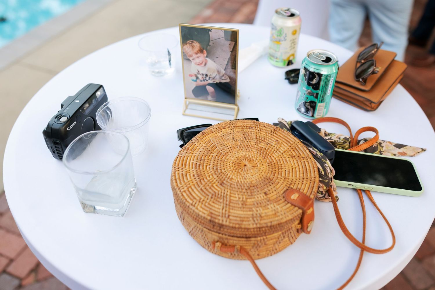 Round rattan bag, film camera, phone, and drinks displayed on white table at outdoor party event.