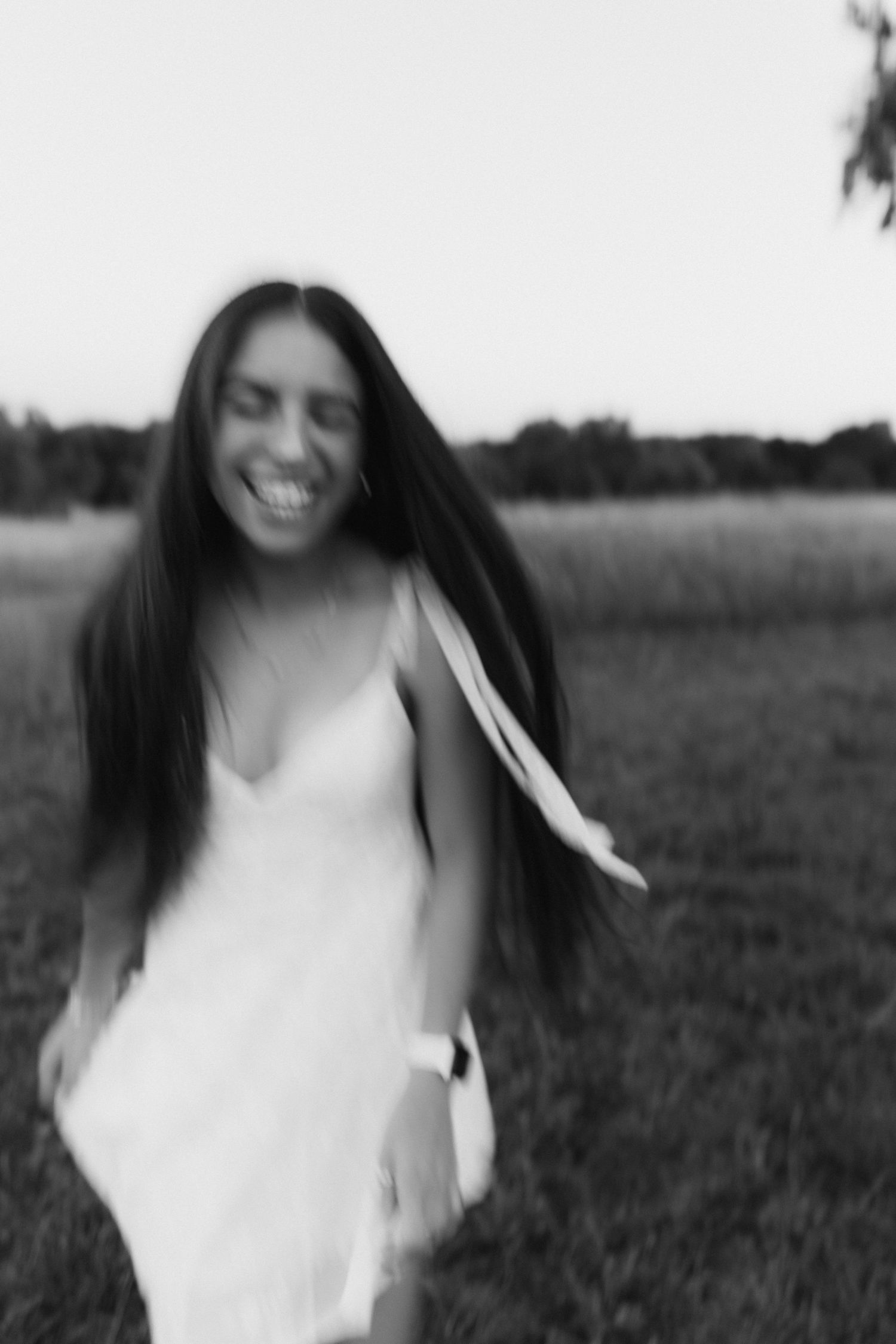 Joyful woman in white dress laughs with hair flowing in motion blur in black and white field photo.