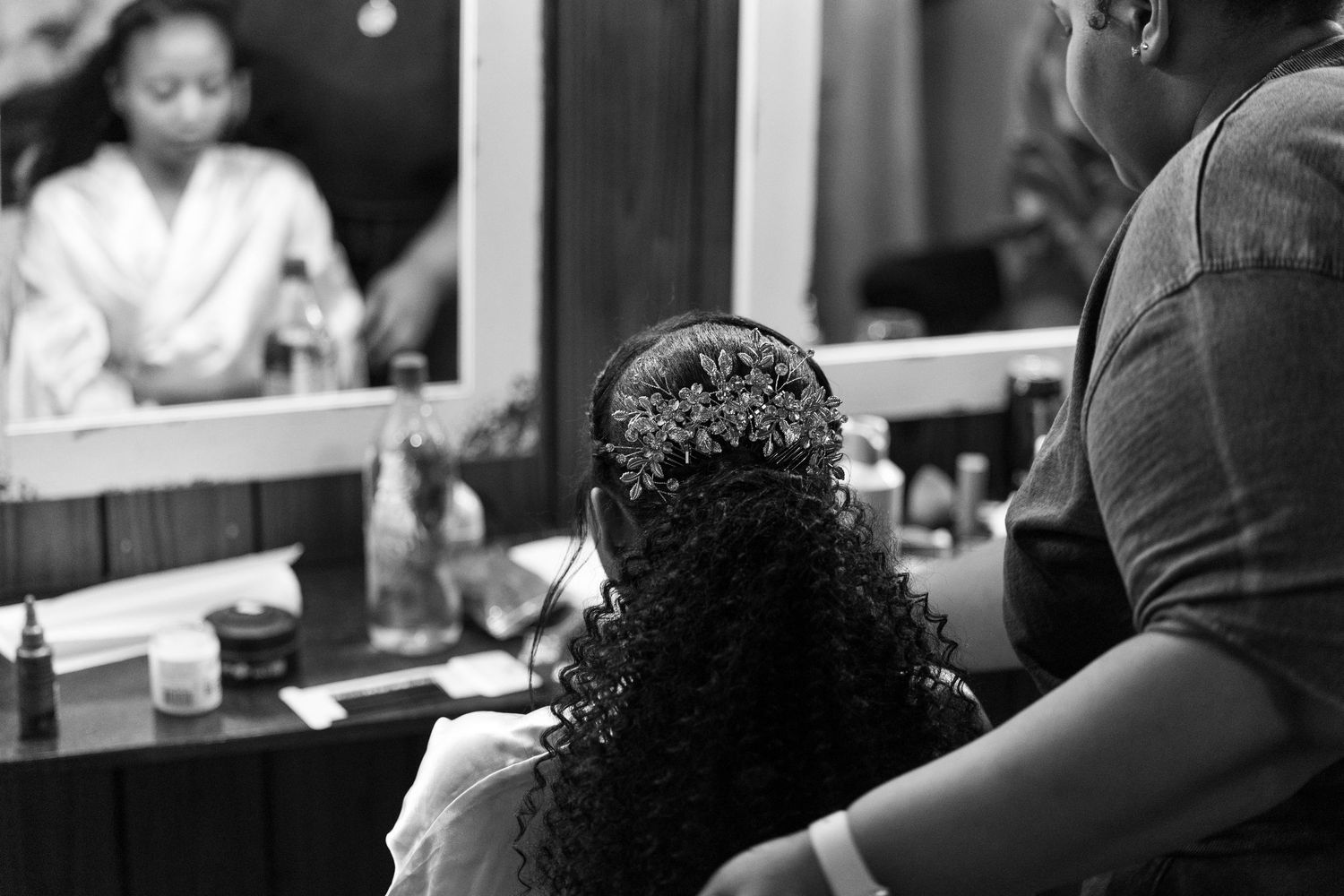 Black and white photo of a woman having her curly hair styled at a vanity mirror backstage.