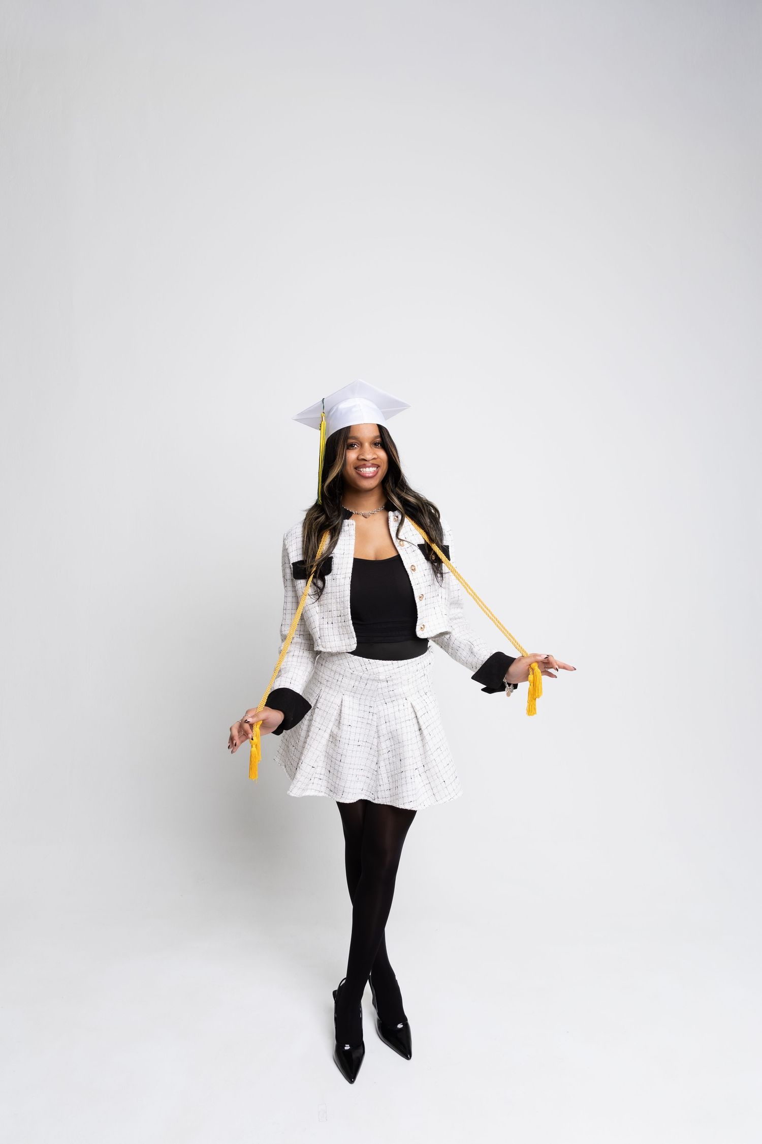 Smiling graduate in white cap and blazer with gold tassels, black skirt and tights, posing on white background.