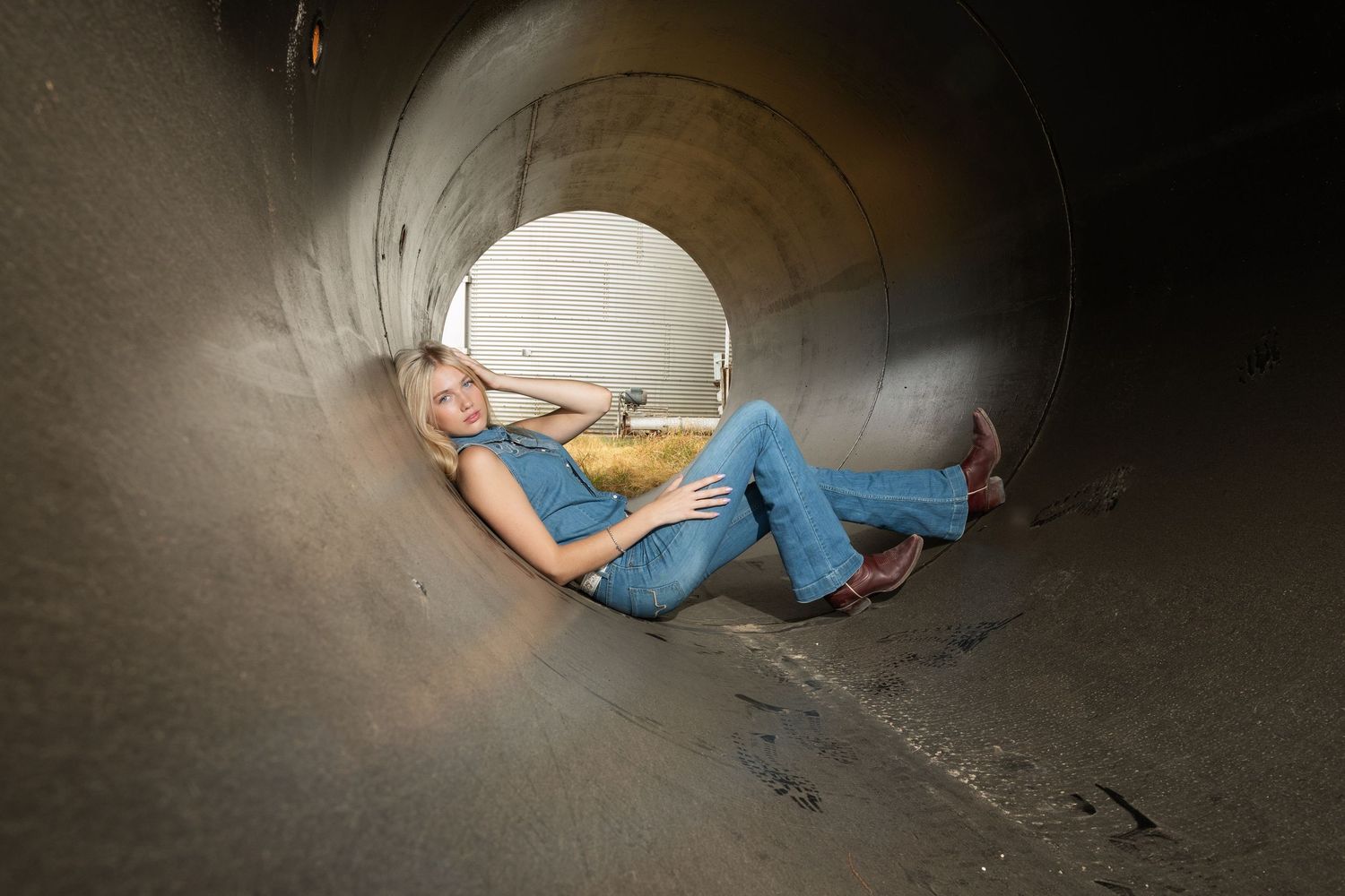 Blonde woman in jeans posing inside a large industrial metal tunnel or pipe.