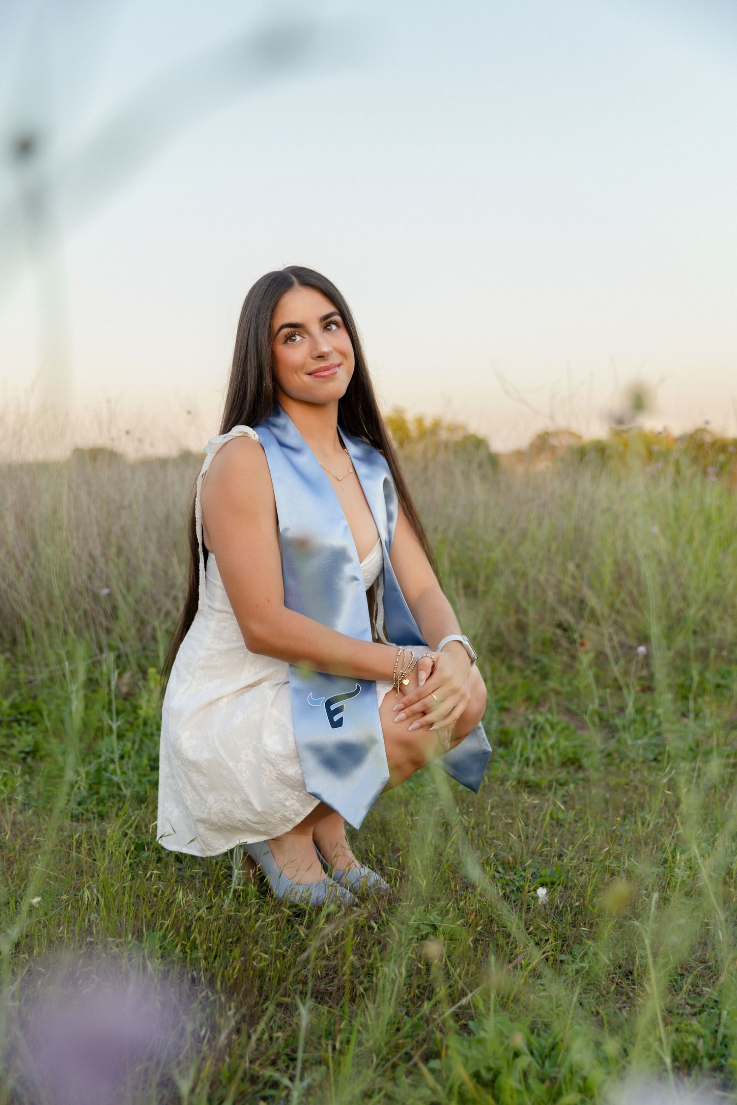 Young woman in white dress with blue graduation sash kneeling in a field at sunset for senior photos.