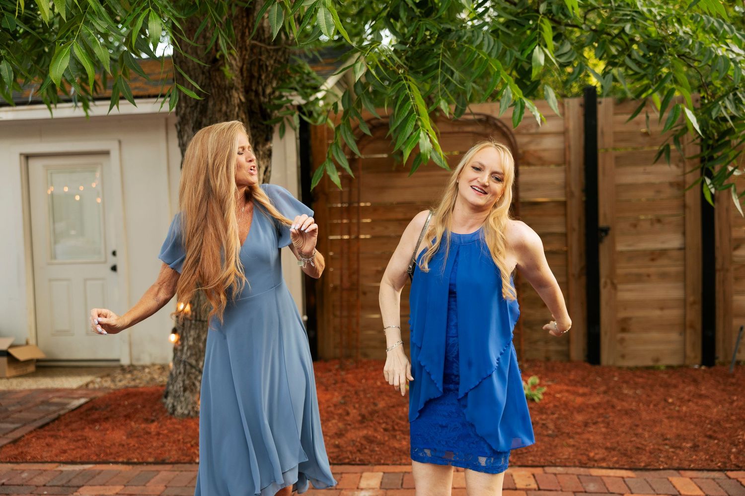 Two women in blue dresses dancing joyfully at an outdoor wedding celebration.