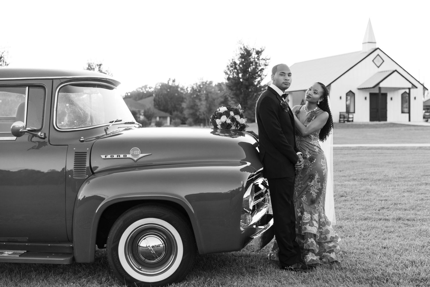 Black and white photo of a couple posing by a vintage pickup truck outside a white church on their wedding day.