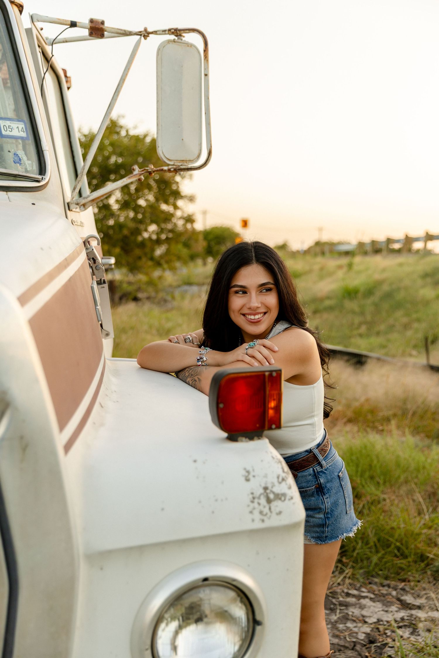 Young woman with tattoos smiles leaning on a vintage white van at sunset in a rural field.
