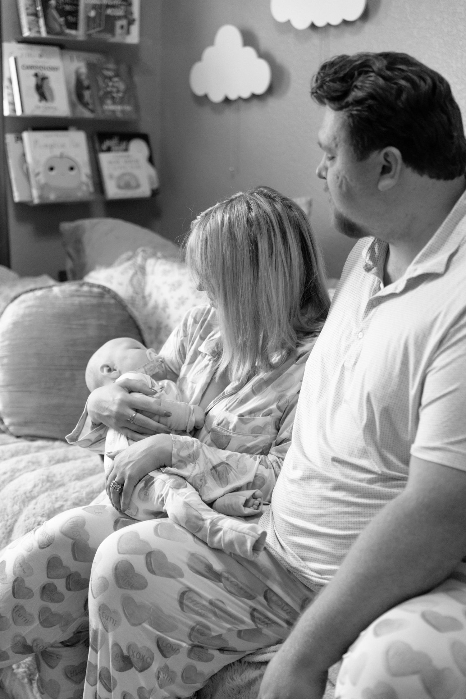 Black and white photo of new parents sitting on a bed in a nursery as mother bottle feeds their newborn baby.