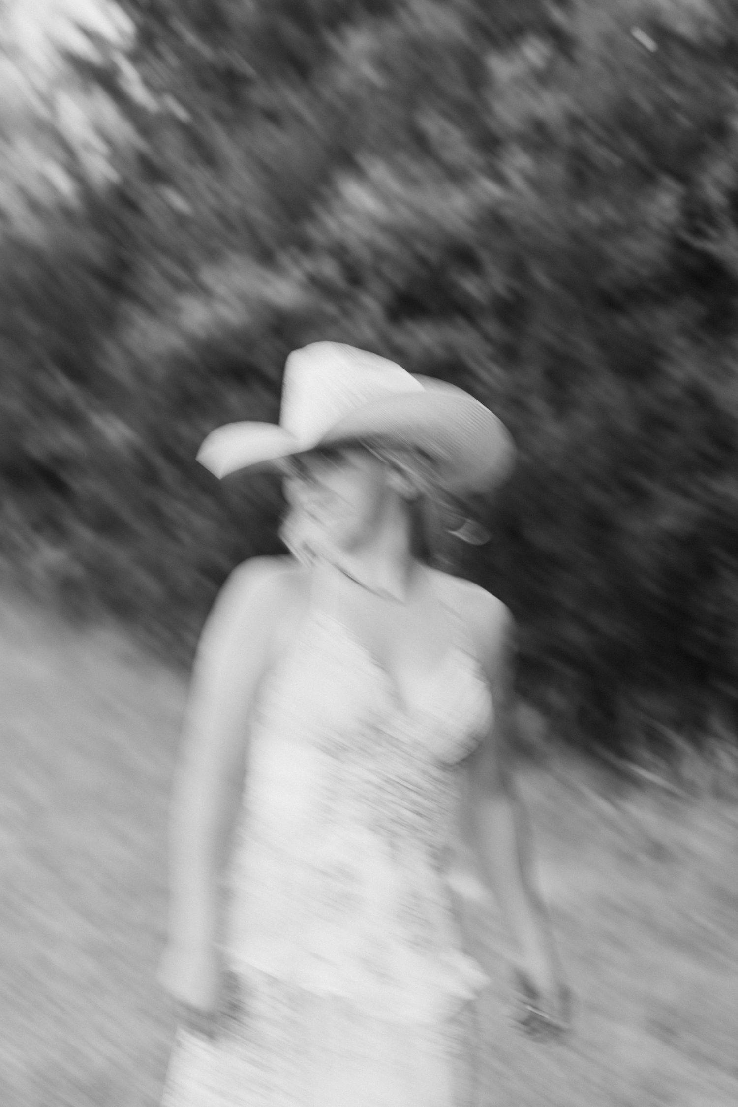 Blurred black and white photo of a woman in a cowboy hat and floral dress walking outdoors.