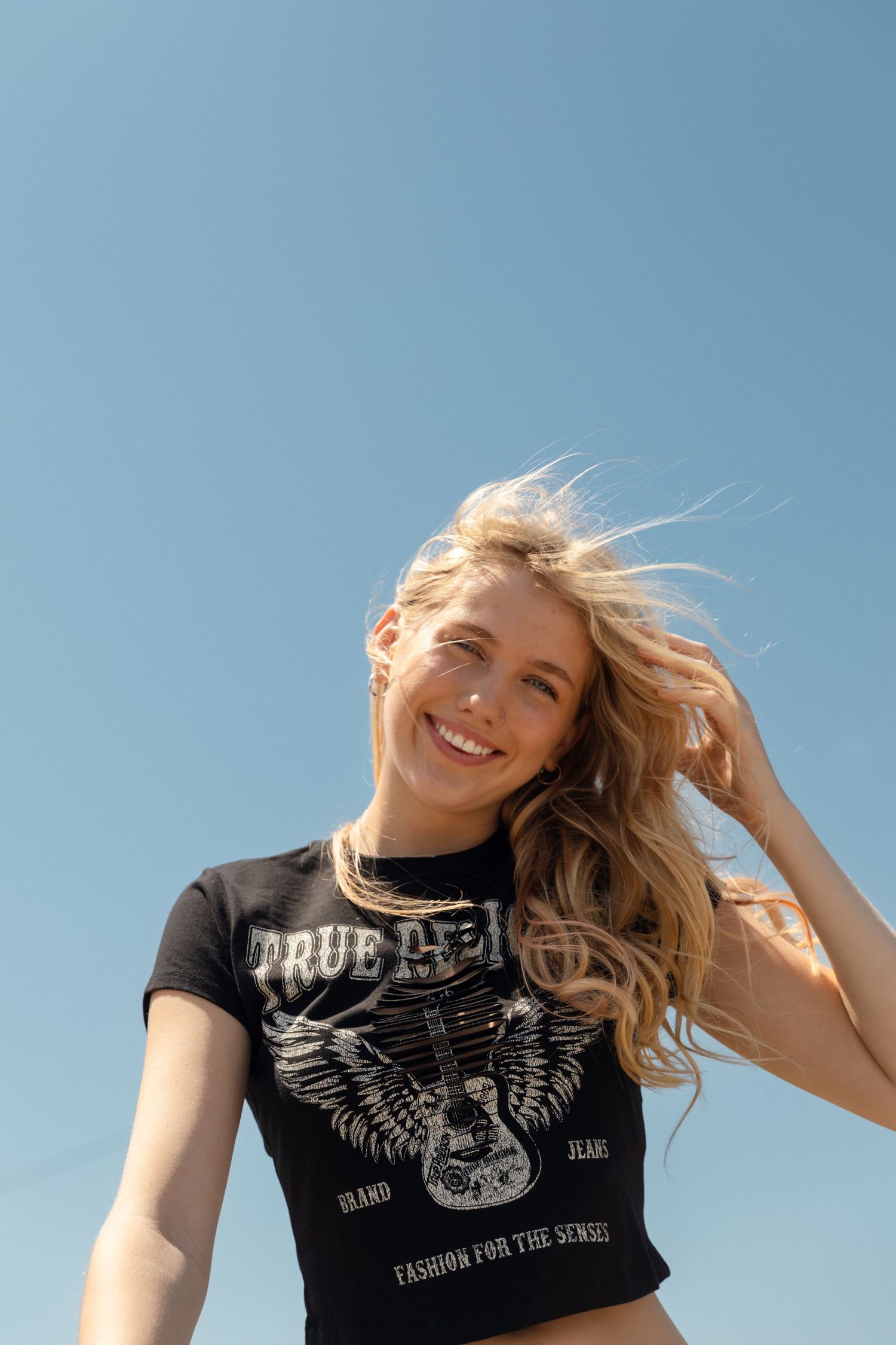 Smiling blonde woman in black True Religion graphic tee with wind-blown hair against clear blue sky.
