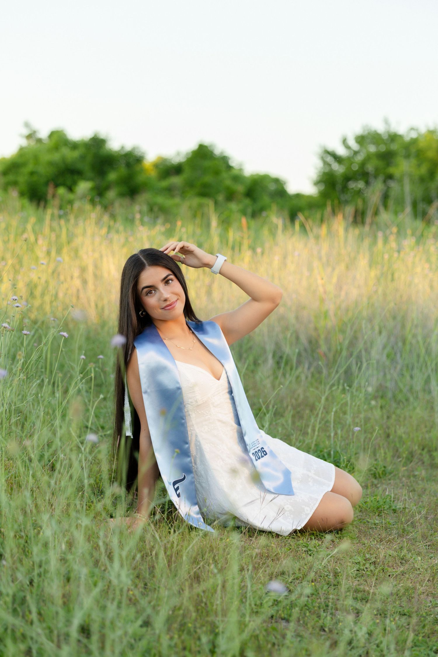 Young woman in white dress and blue graduation stole sits gracefully in a tall grass field.