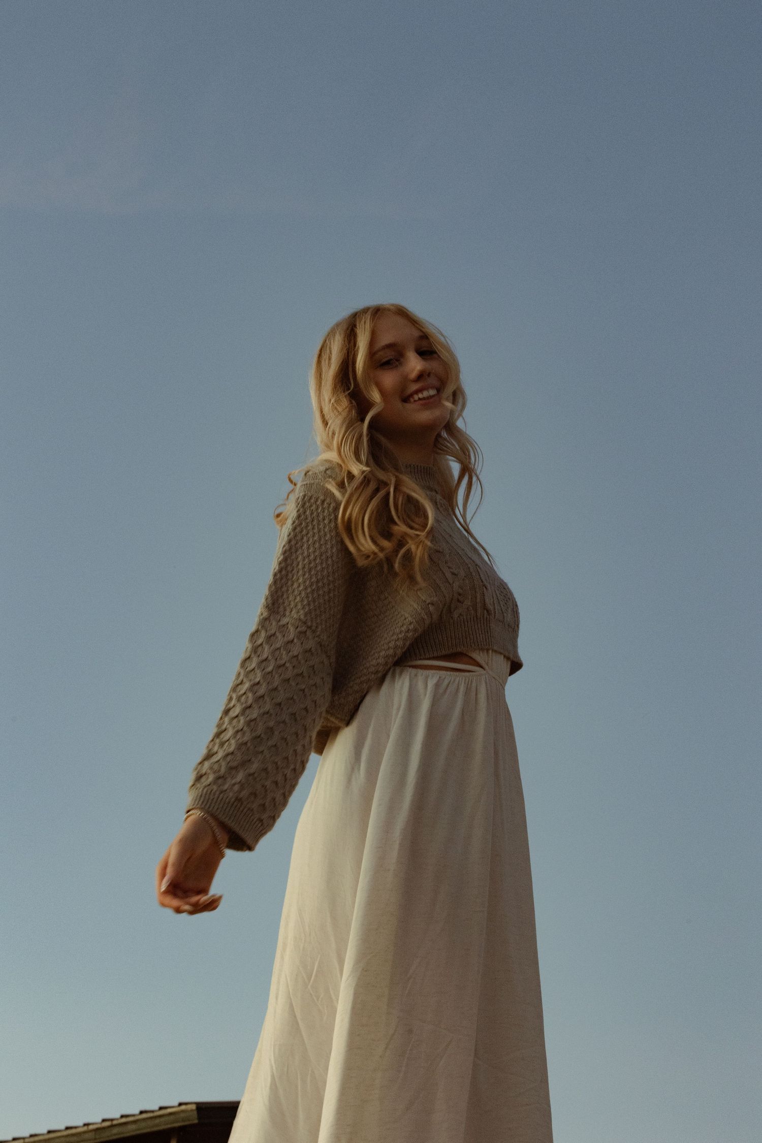 Smiling blonde woman in knit sweater and white skirt photographed from below against blue sky.