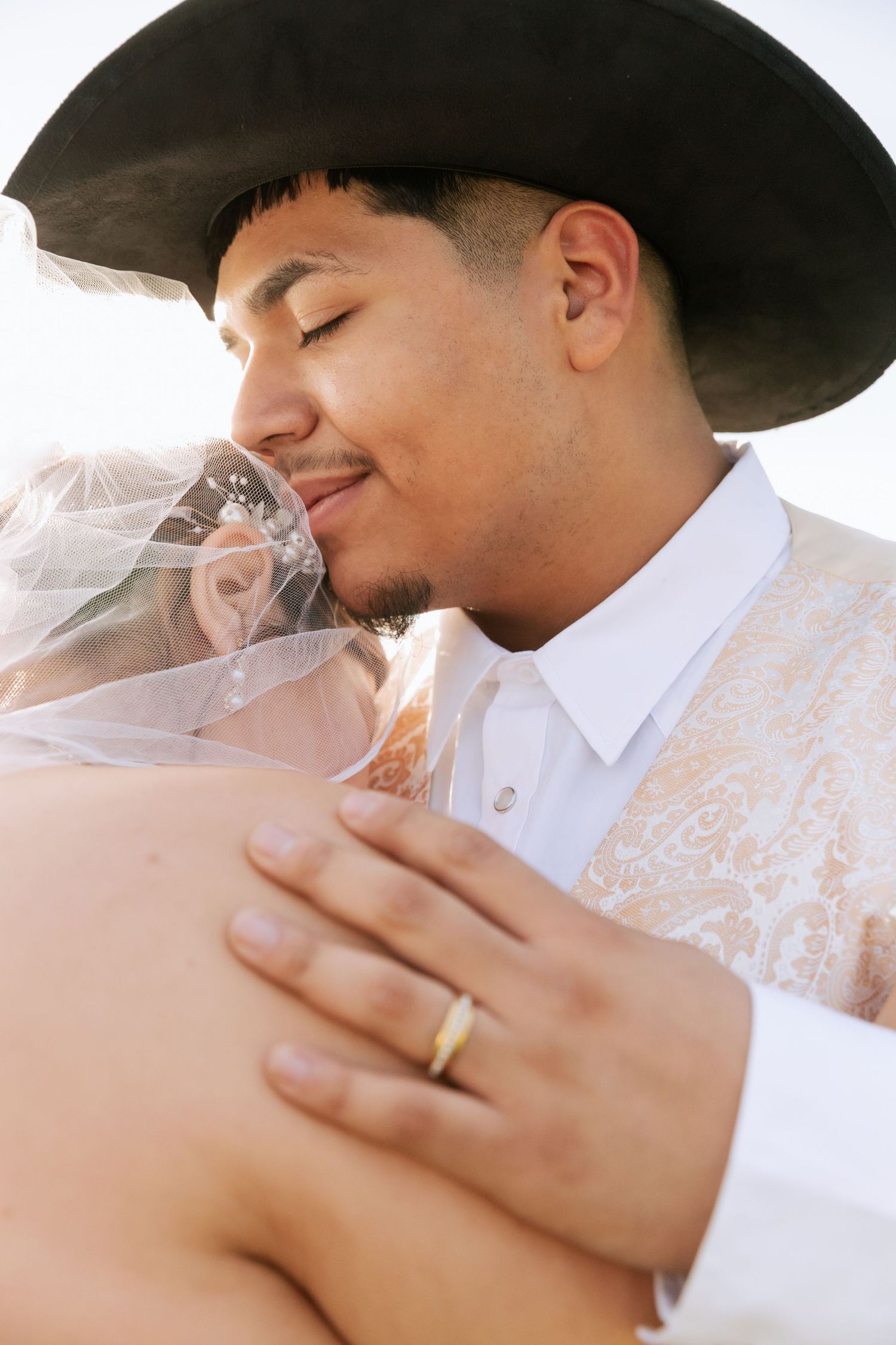 Groom in black cowboy hat tenderly kissing bride through her veil with wedding ring visible on his hand.