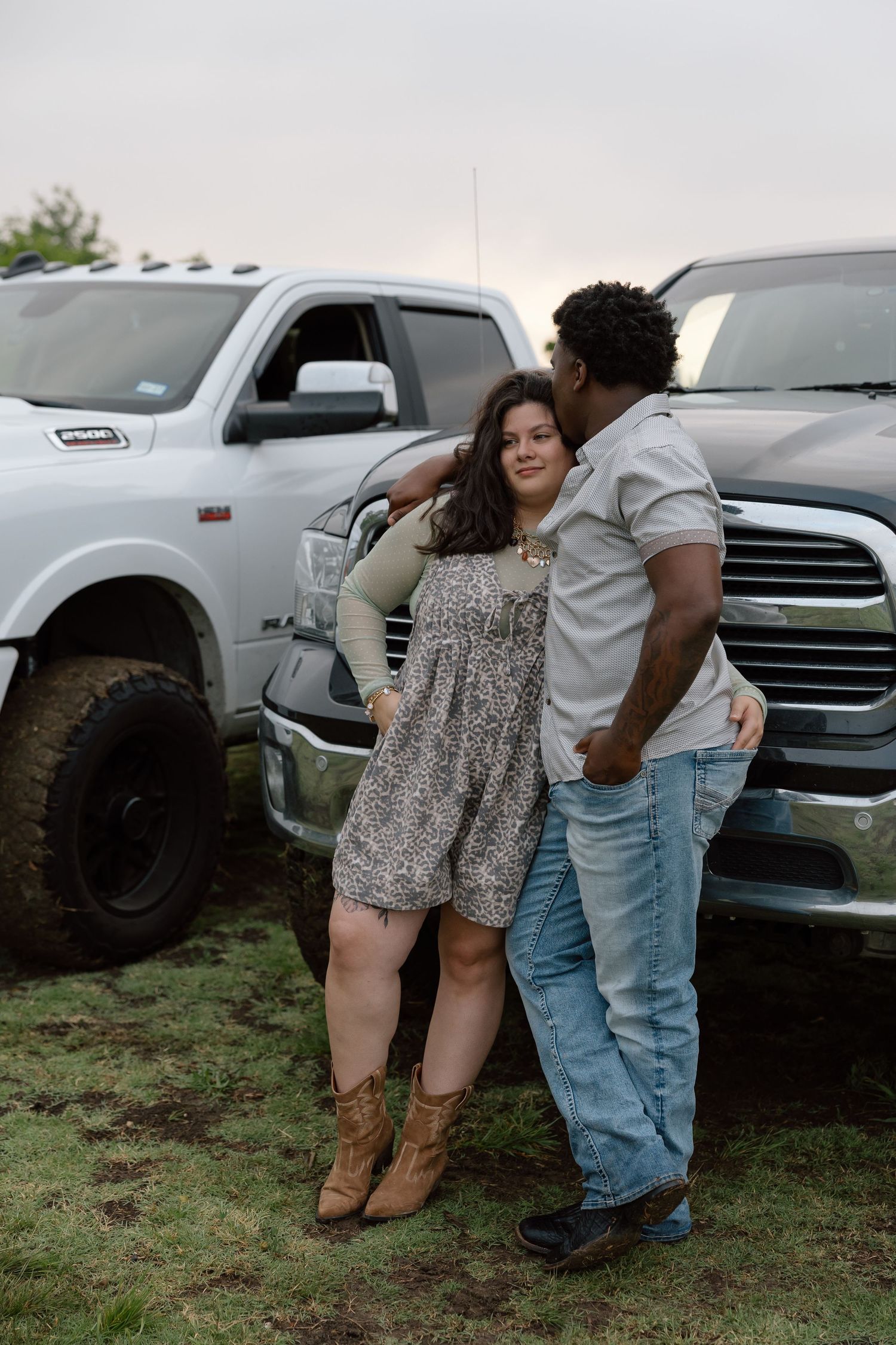 Interracial couple sharing a kiss beside lifted pickup trucks in an outdoor setting at dusk.