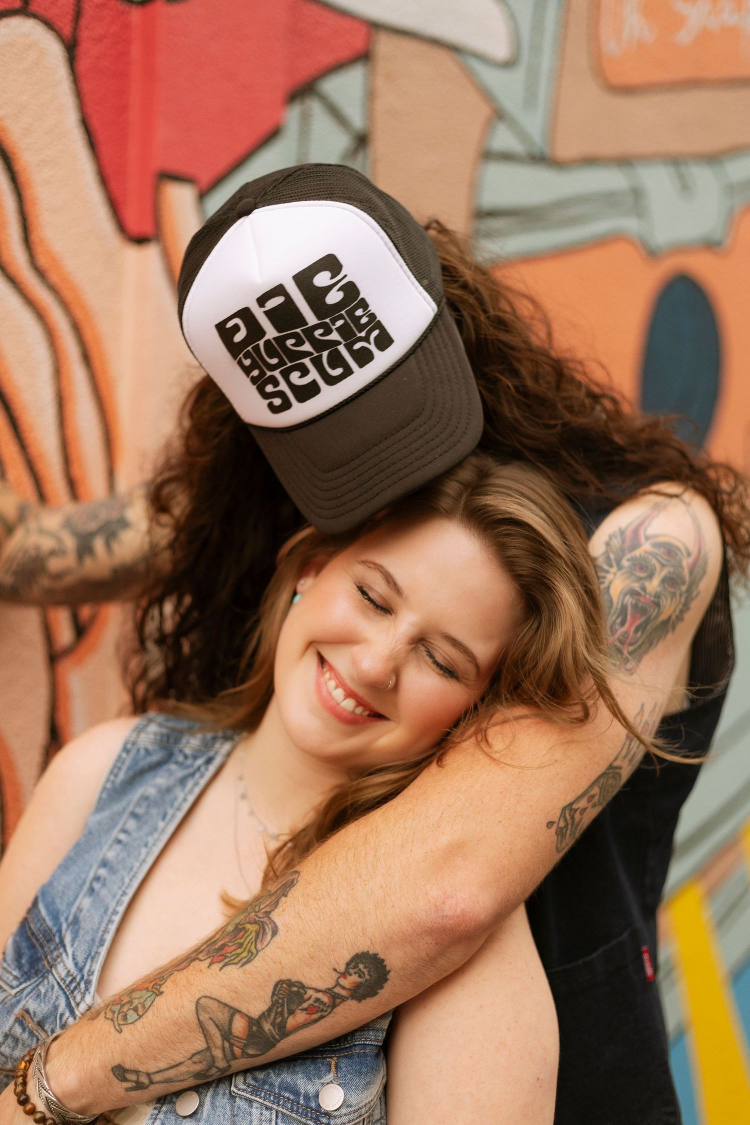 Smiling tattooed woman leans against colorful graffiti wall while partner places hat on her head.