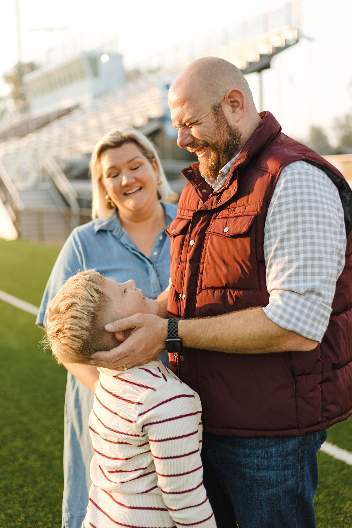 A happy family of three shares a joyful moment on a green sports field, with stadium bleachers in the background.