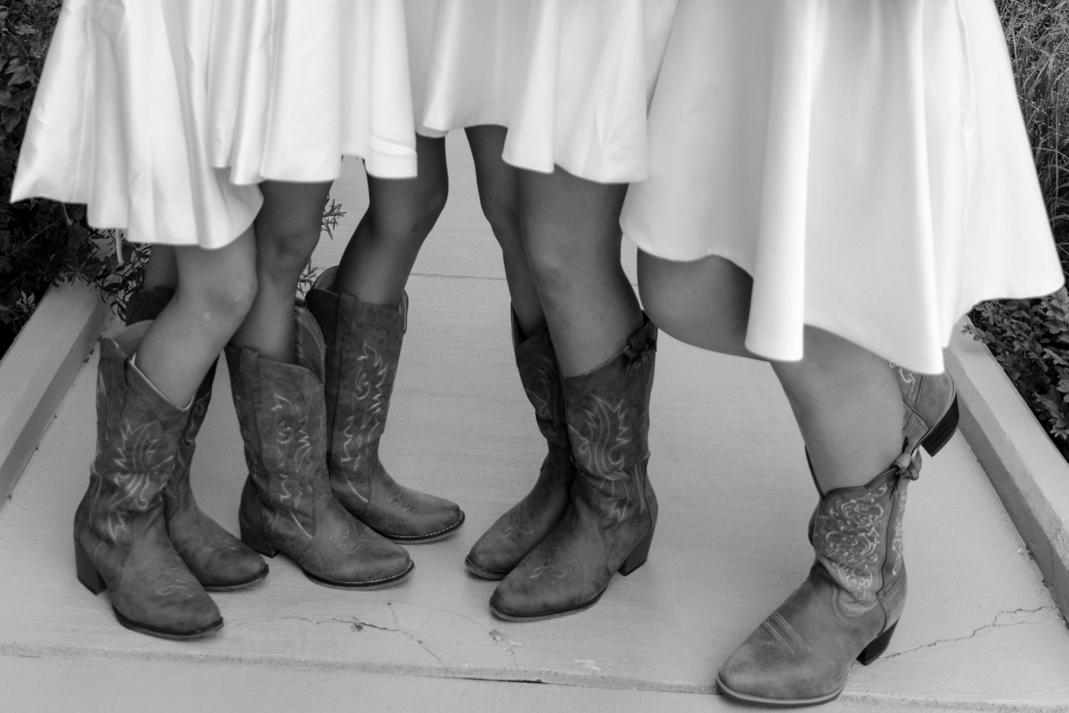 Black and white photo of bridesmaids in white dresses wearing matching ornate cowboy boots at a wedding.