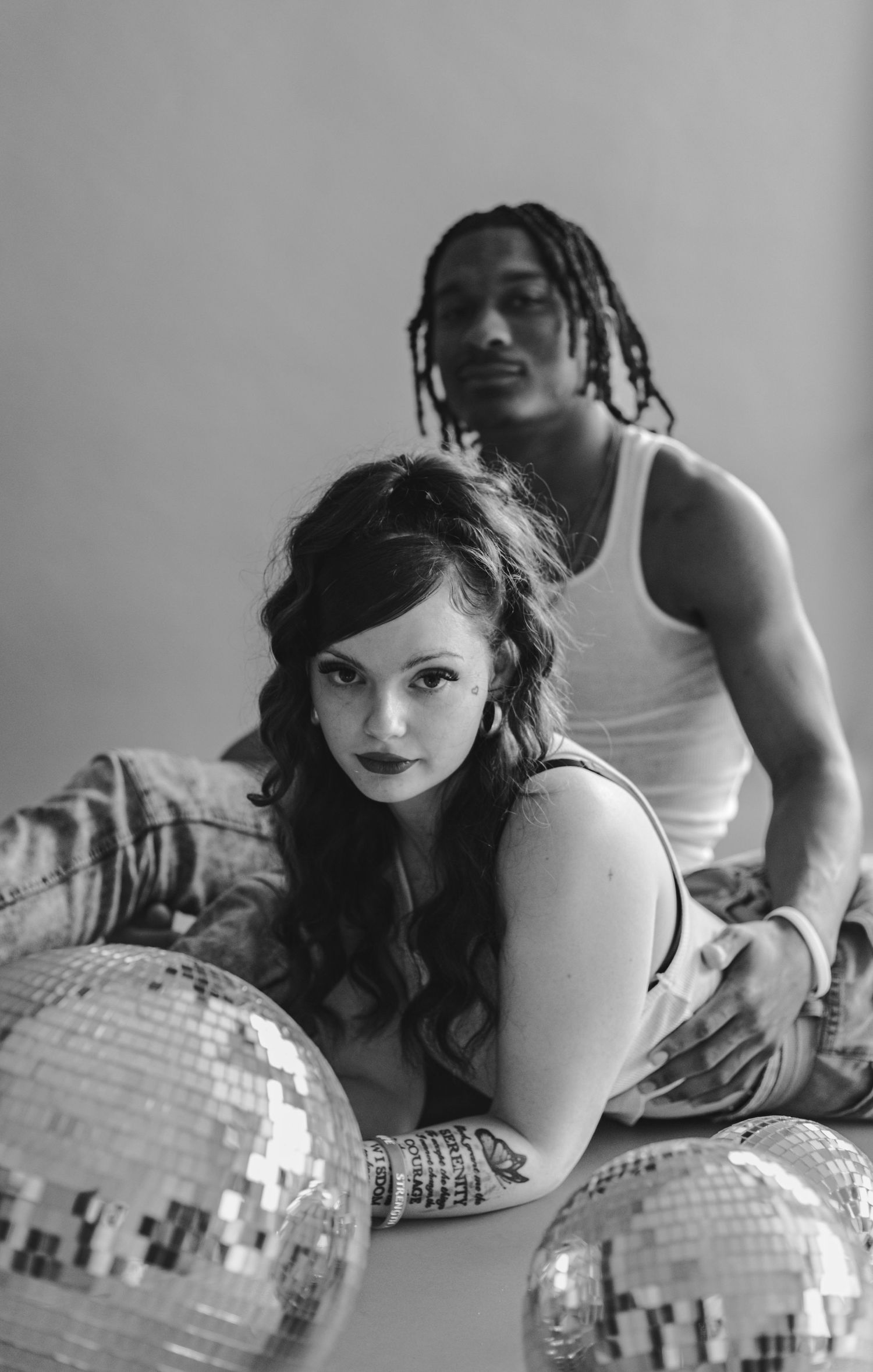 Black and white photo of a couple posing with disco balls, woman in foreground with tattoos, man with dreads behind her.
