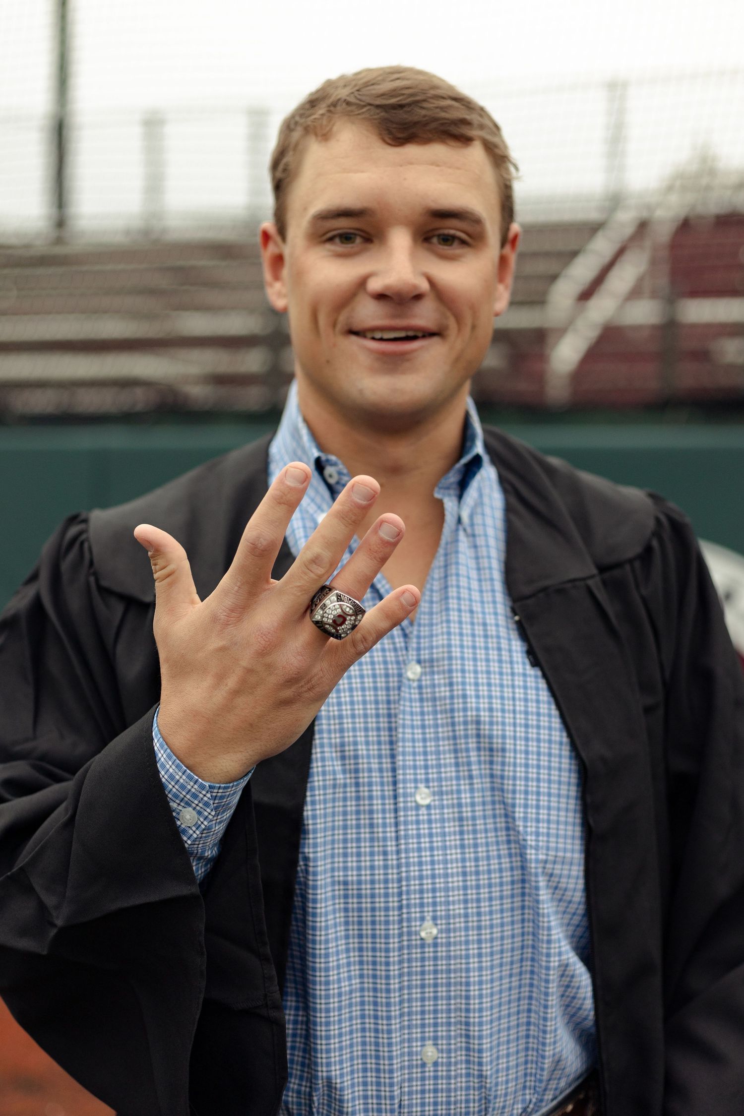 Smiling young man in graduation gown displaying a large championship ring at a sports stadium.
