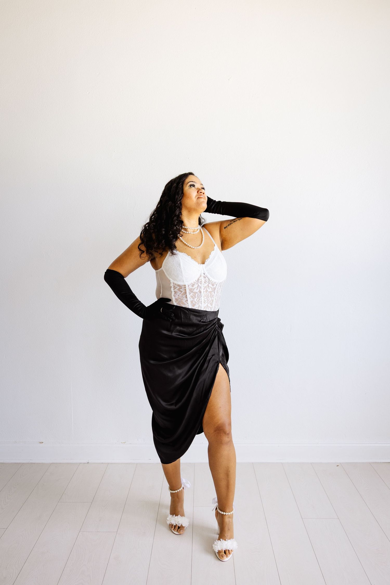 Woman posing in white lace corset top, black satin wrap skirt, and white heels with black gloves.