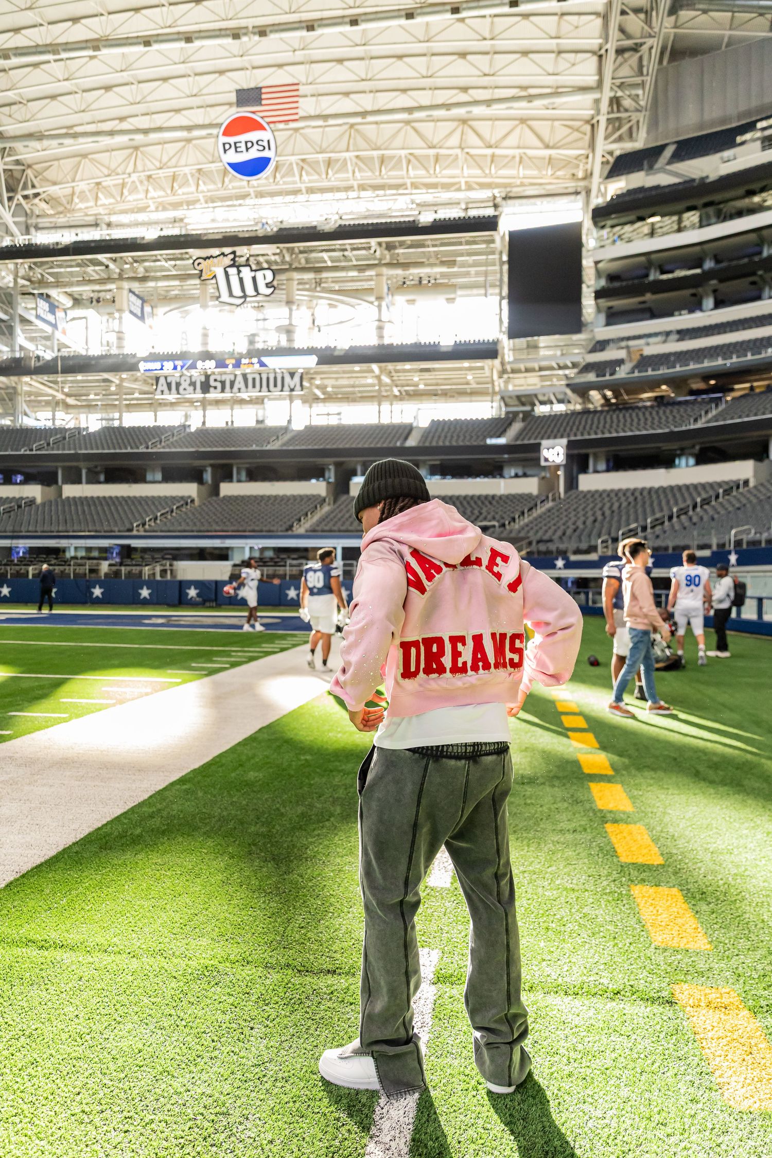 Person wearing pink Dreams hoodie stands on the field at AT&T Stadium during a football event.