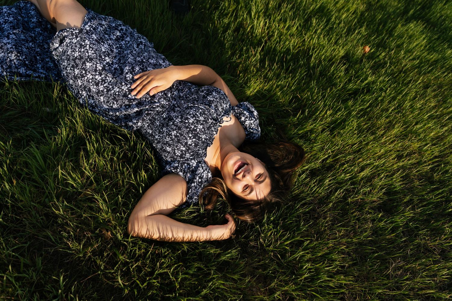 Young woman in floral dress lies peacefully in lush green grass during golden hour sunlight.
