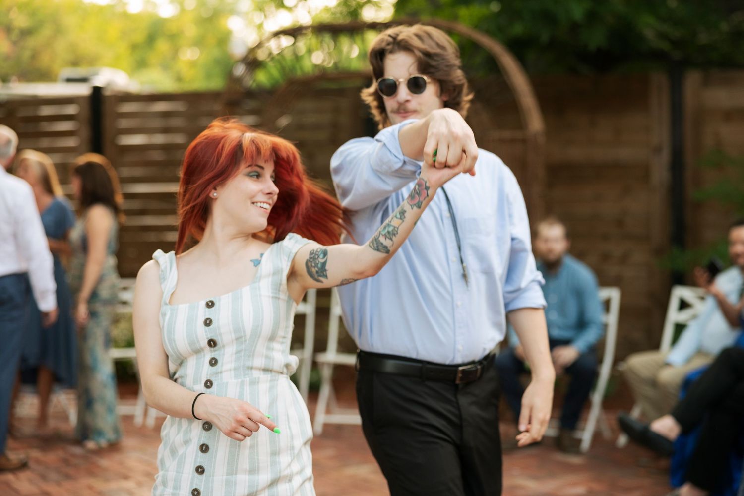 Red-haired woman in floral dress dances with tattooed man in blue shirt at outdoor wedding reception.