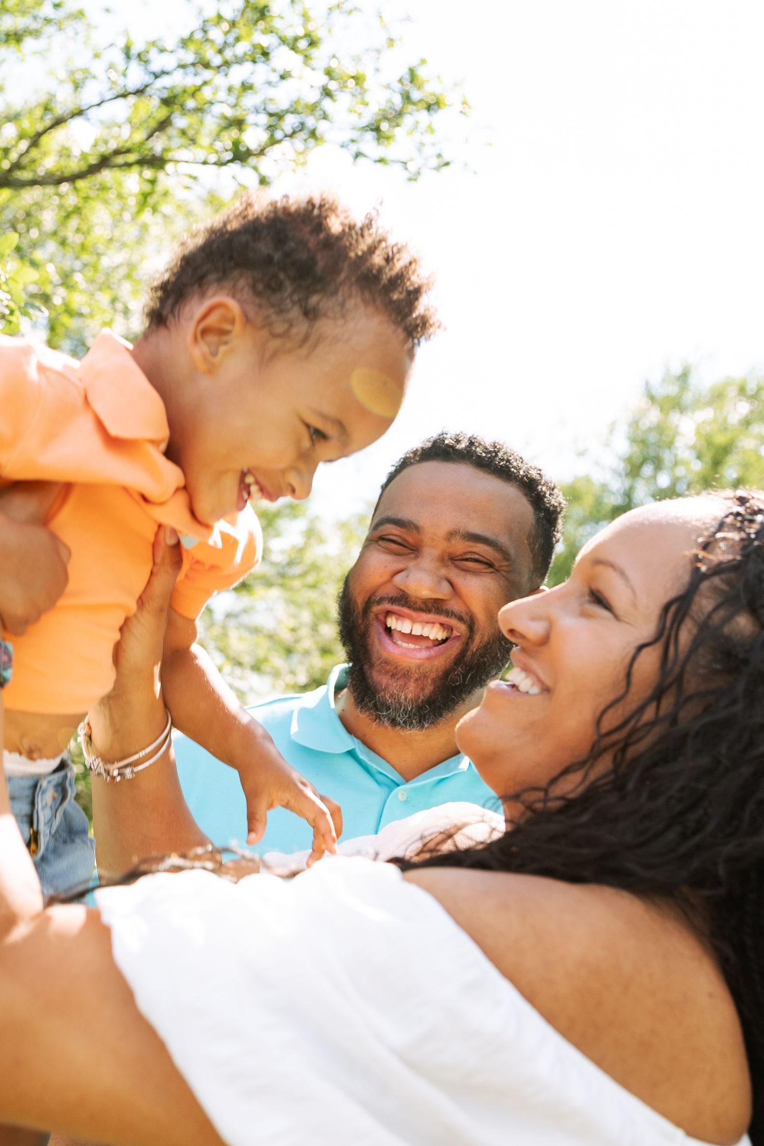 Joyful Black family outdoors, parents laughing as they lift smiling baby in orange outfit in sunny park.