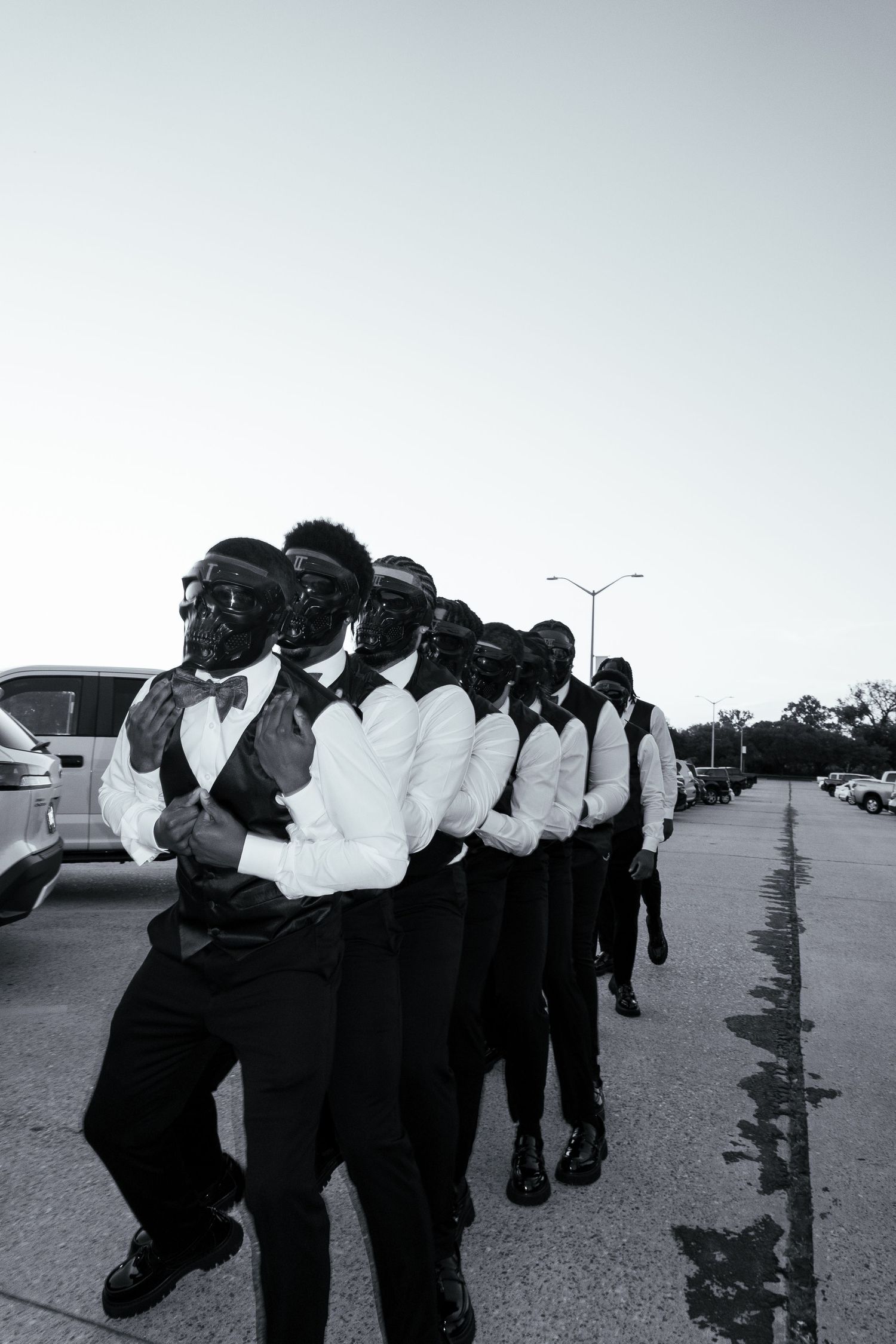 Black and white photo of a group of men in formal attire with masks marching in a line outdoors.