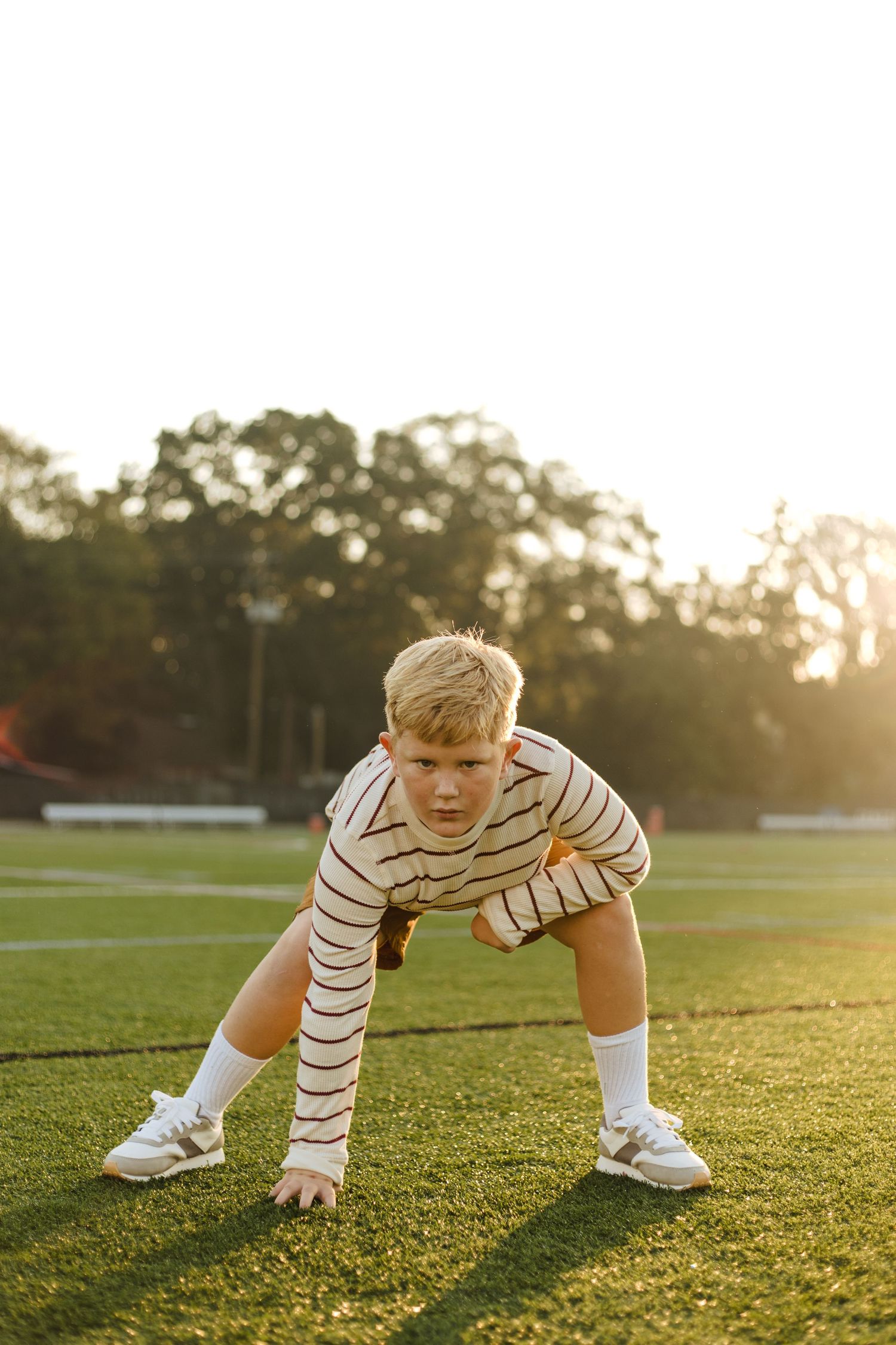 Focused blonde boy in striped shirt in athletic stance on green soccer field at golden hour.