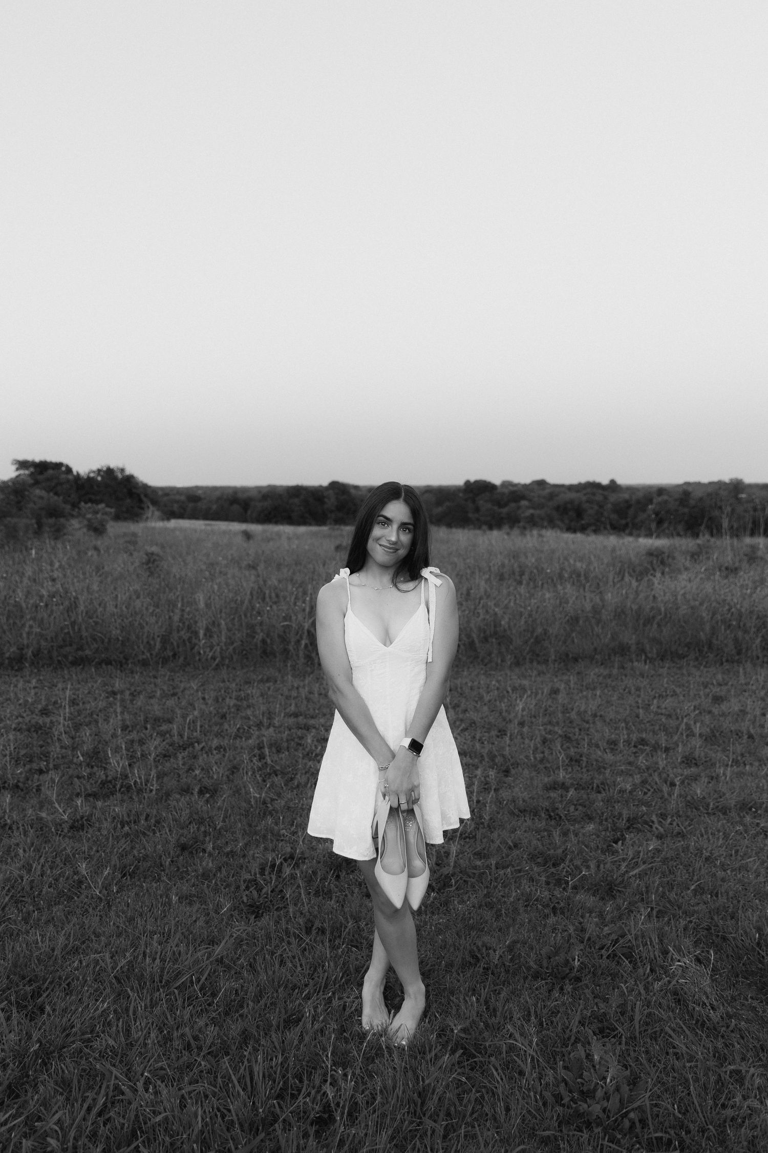 Young woman in white sundress stands barefoot in open field at dusk in black and white photo.