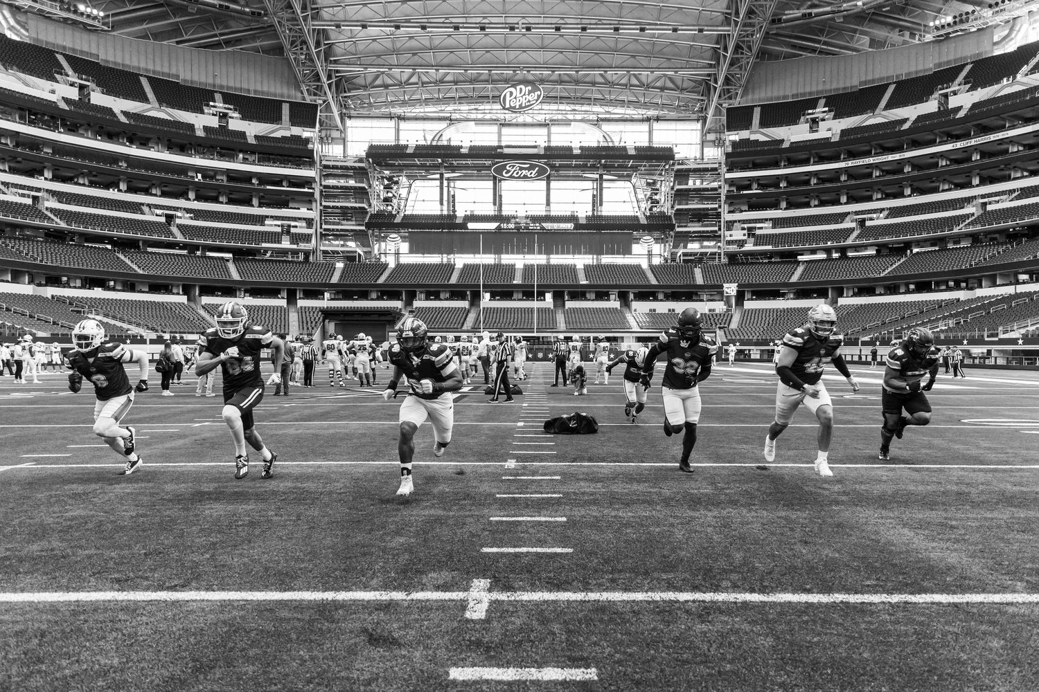 Black and white photo of football players warming up on the field inside a large NFL stadium.