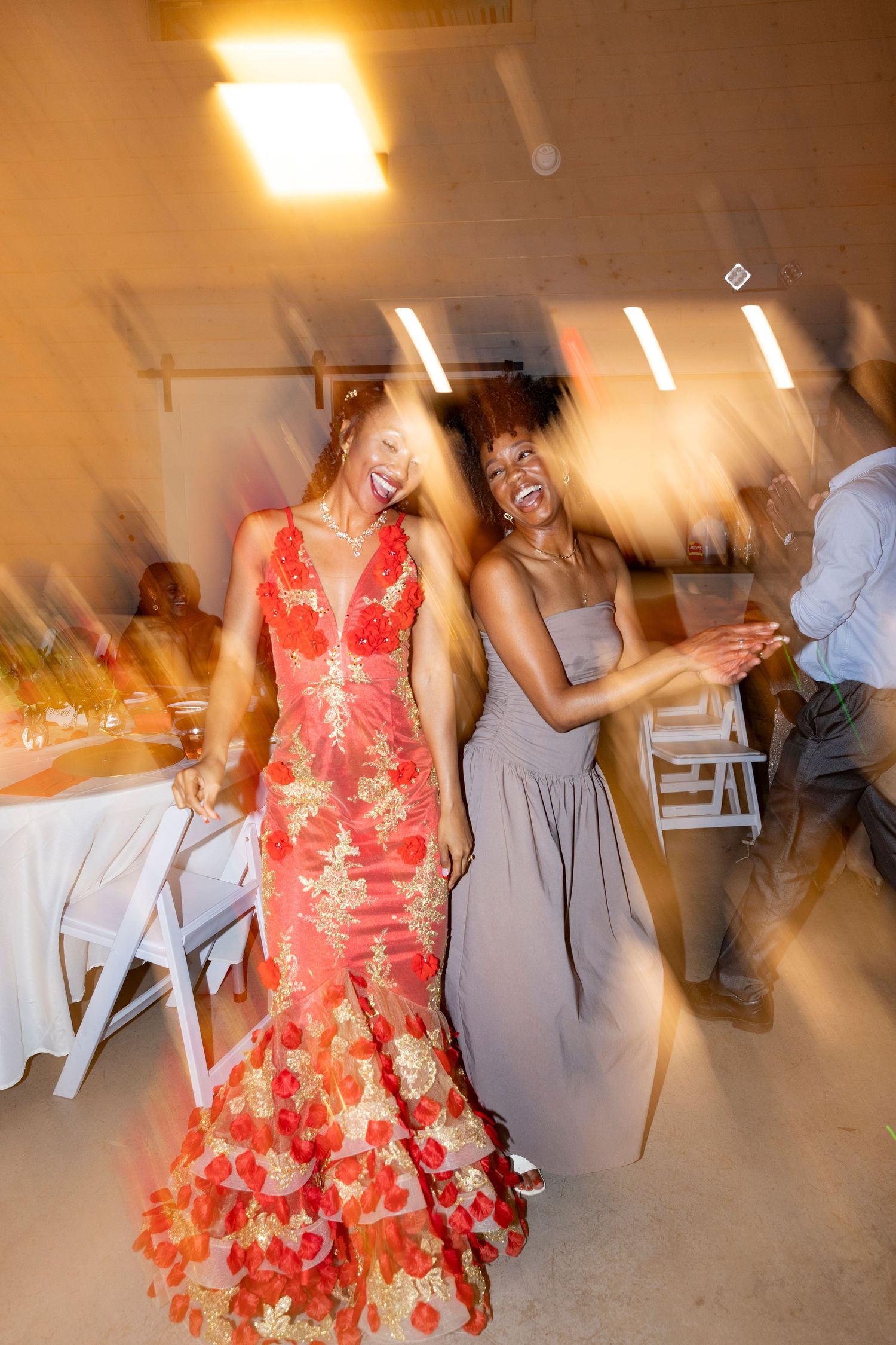 Motion-blurred photo of a bride in a red floral gown dancing joyfully with a bridesmaid at a wedding reception.