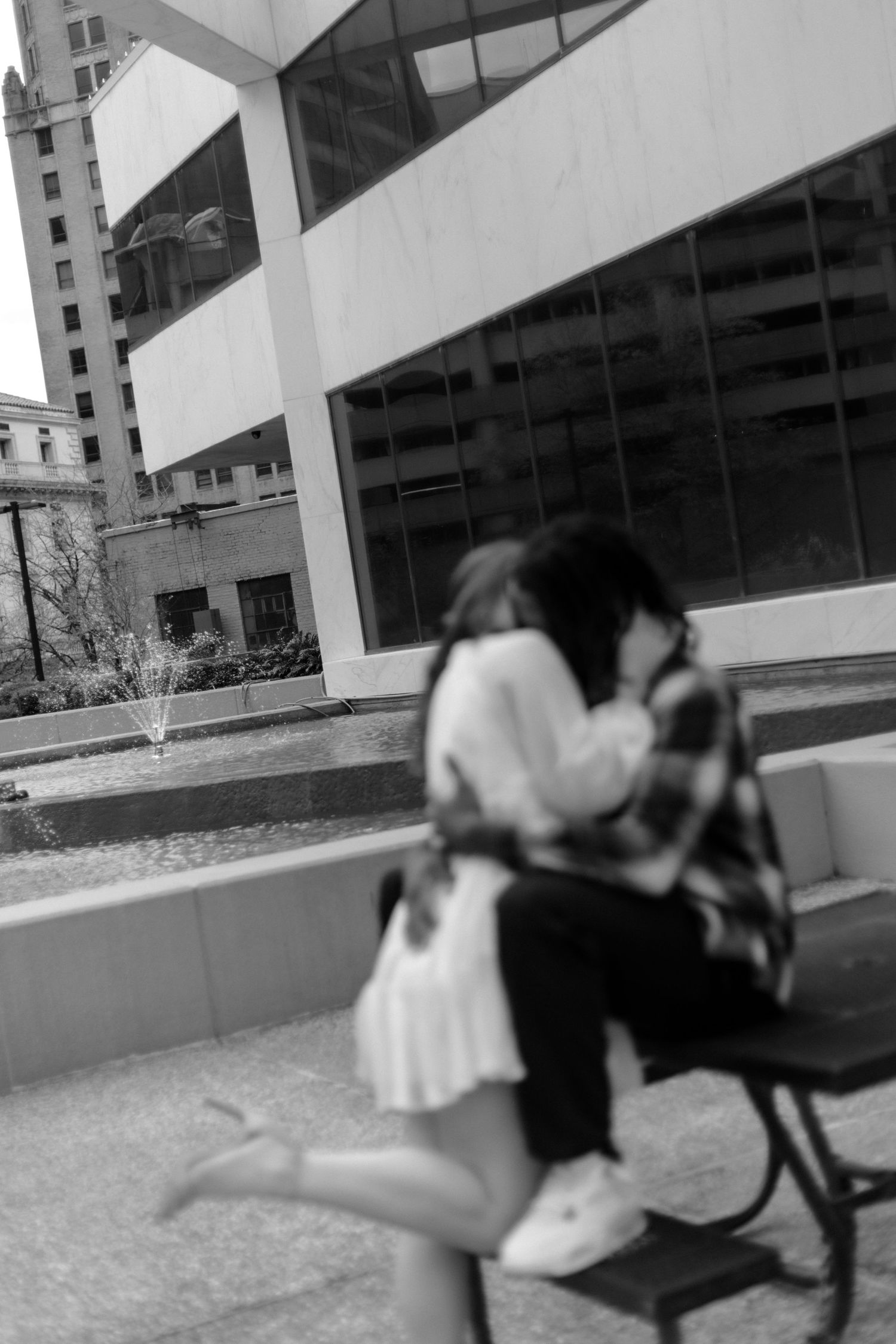 Black and white photo of couple kissing on a bench outside a modern glass building.