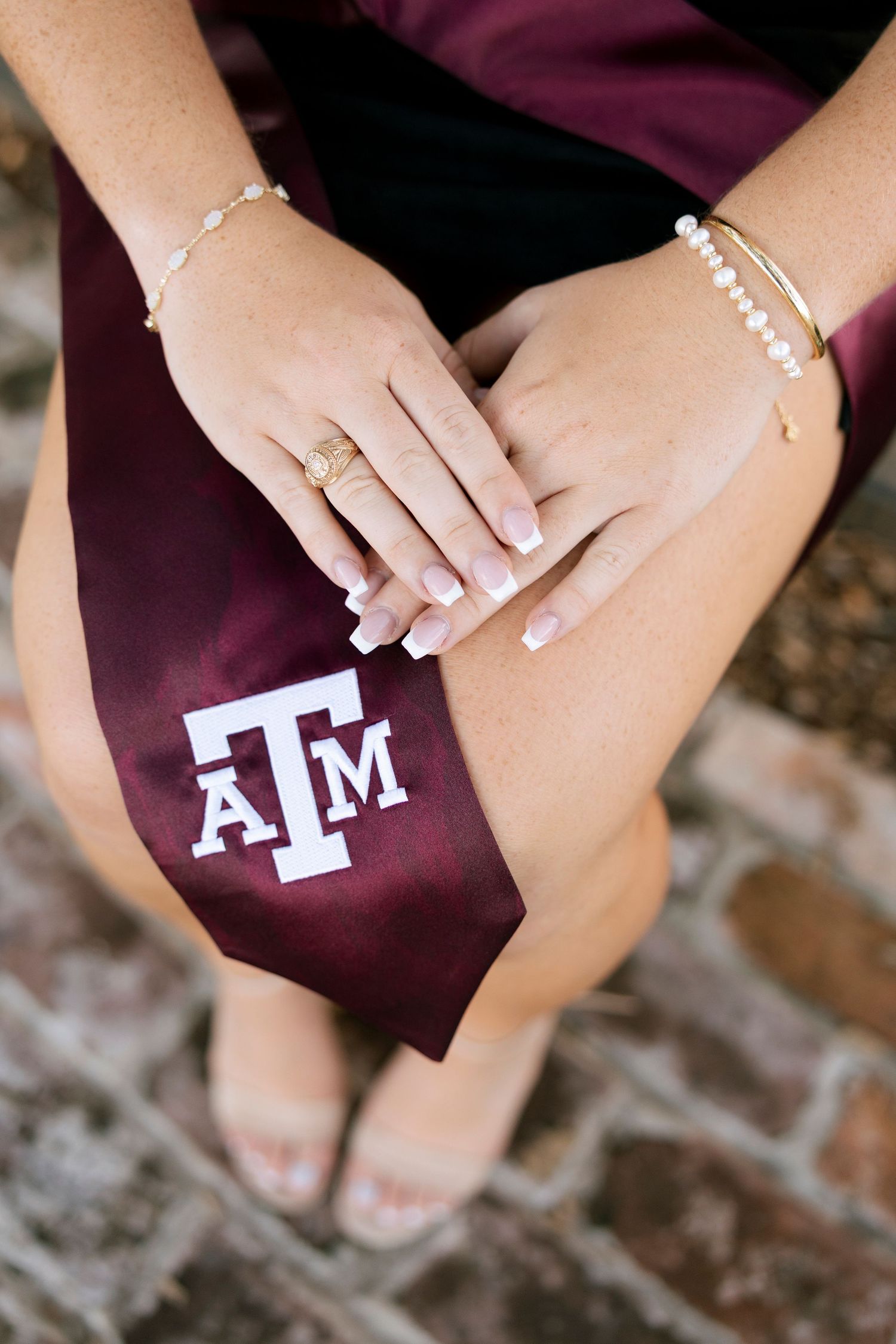 Close-up of hands resting on a maroon Texas A&M graduation sash with pearl and gold bracelets.