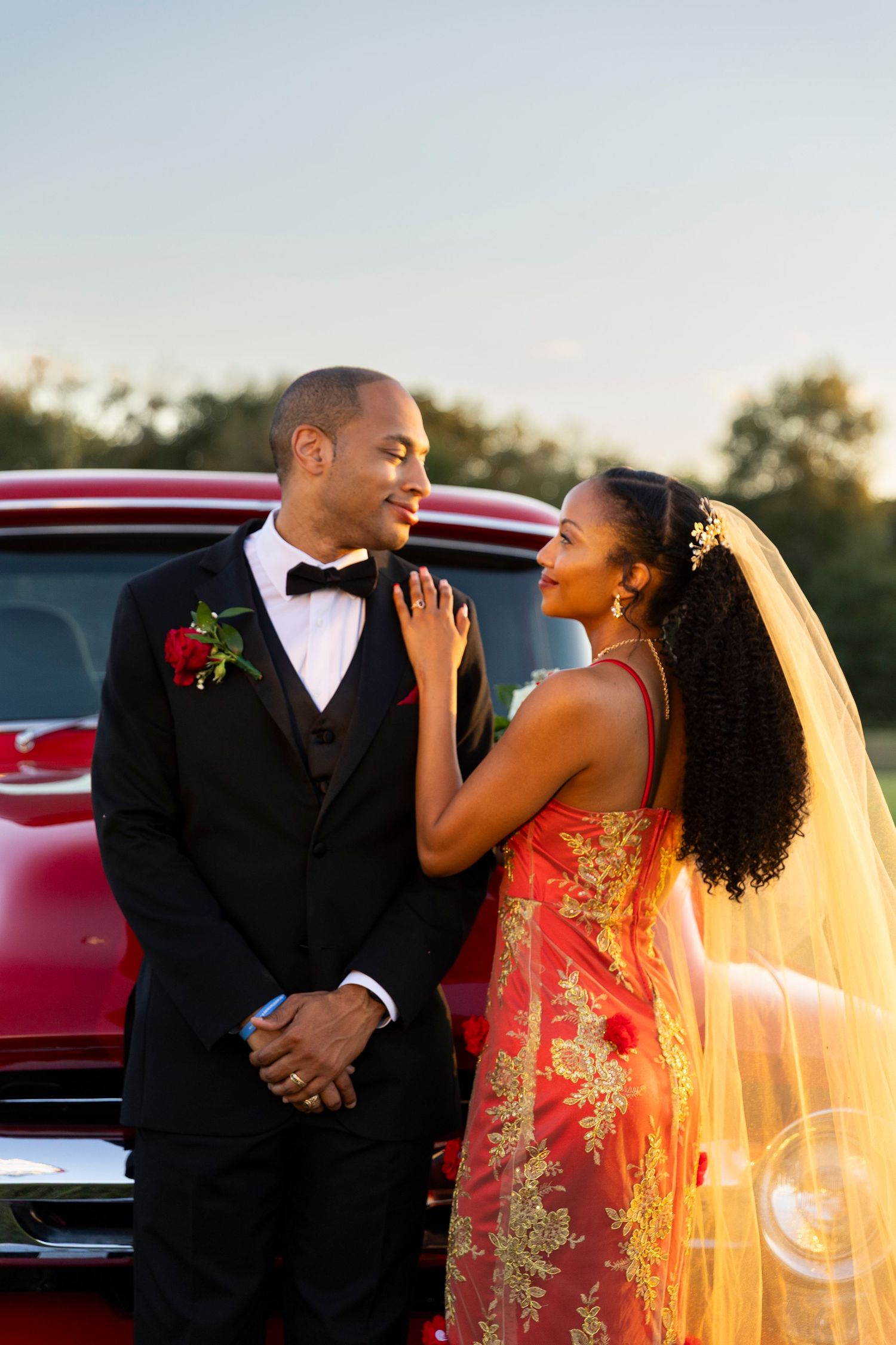 A bride in a red and gold gown and groom in a tuxedo pose by a vintage red truck at sunset.