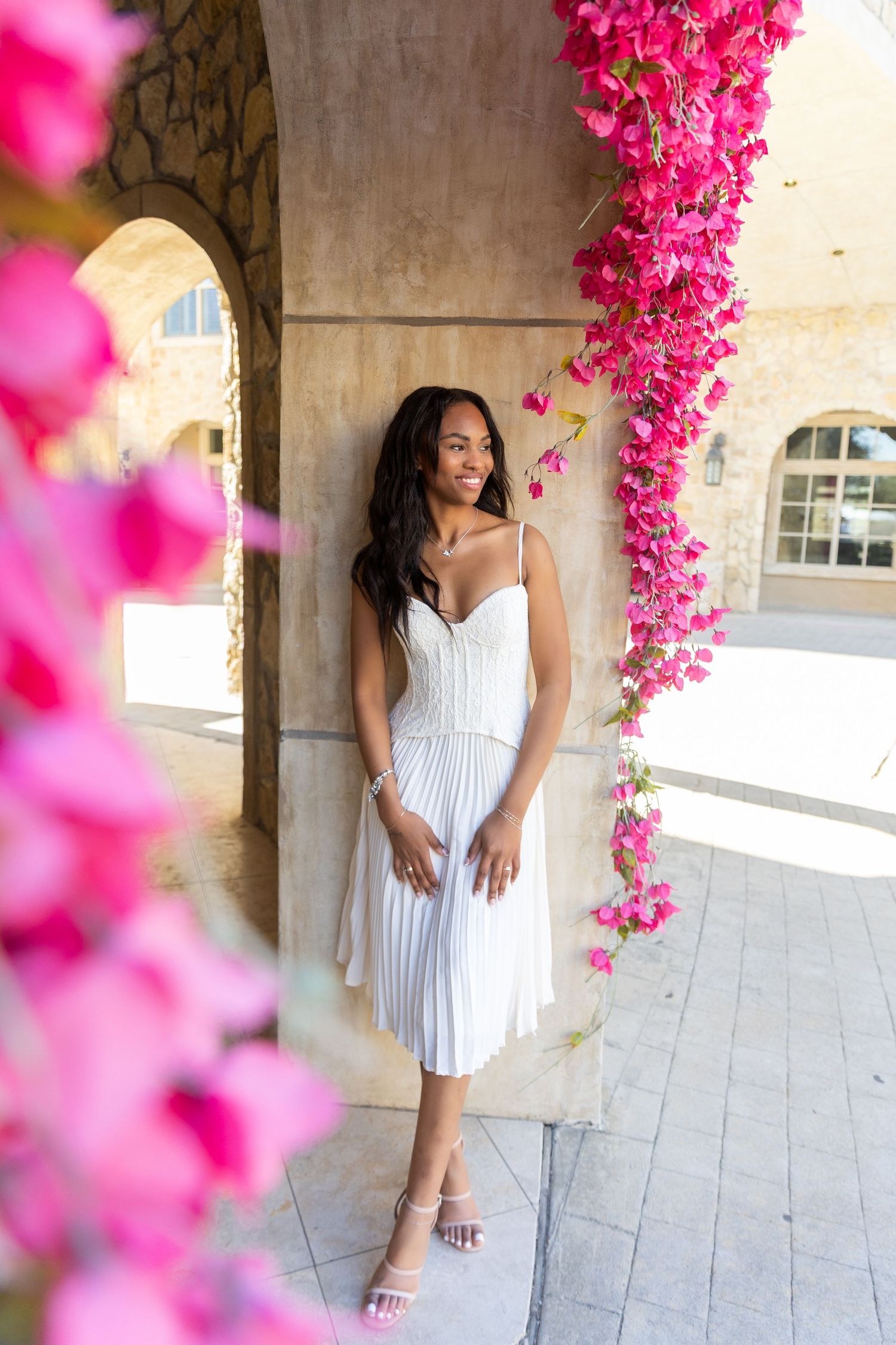 Young Black woman in a white pleated midi dress posing among vibrant pink bougainvillea flowers outdoors.