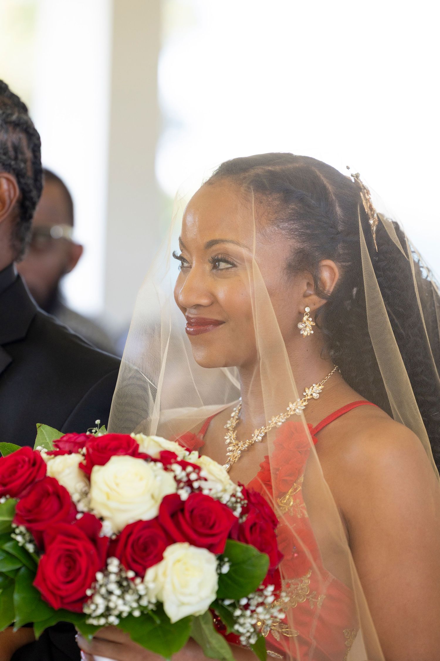 A radiant bride in a red gown holds a bouquet of red and white roses while wearing a veil.