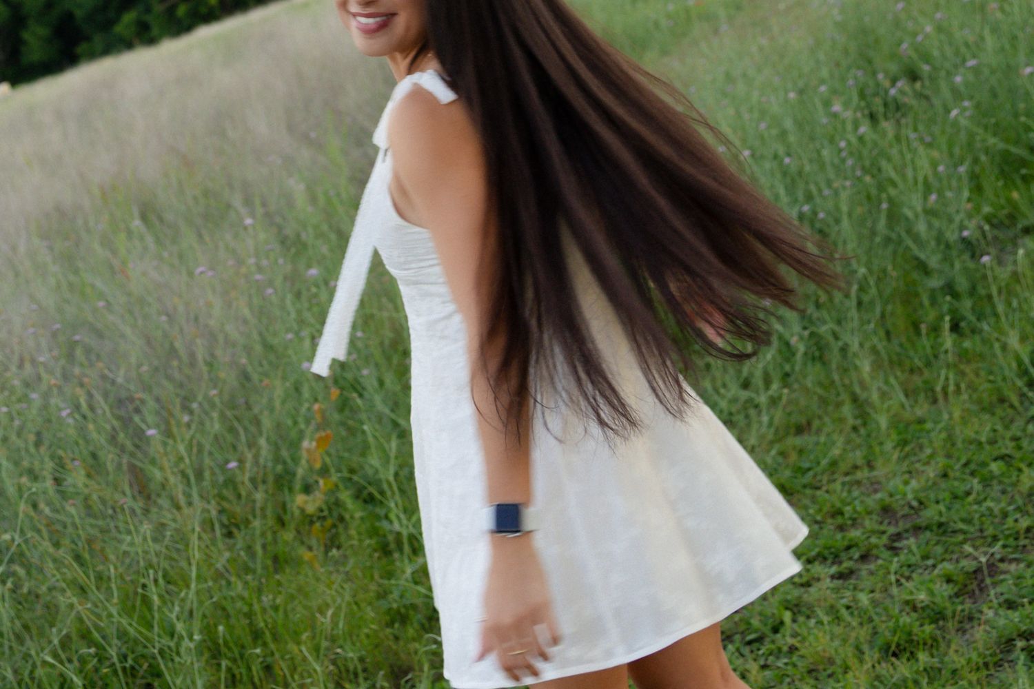 Smiling brunette twirls in a white dress in a lush green meadow, hair flowing in the breeze.