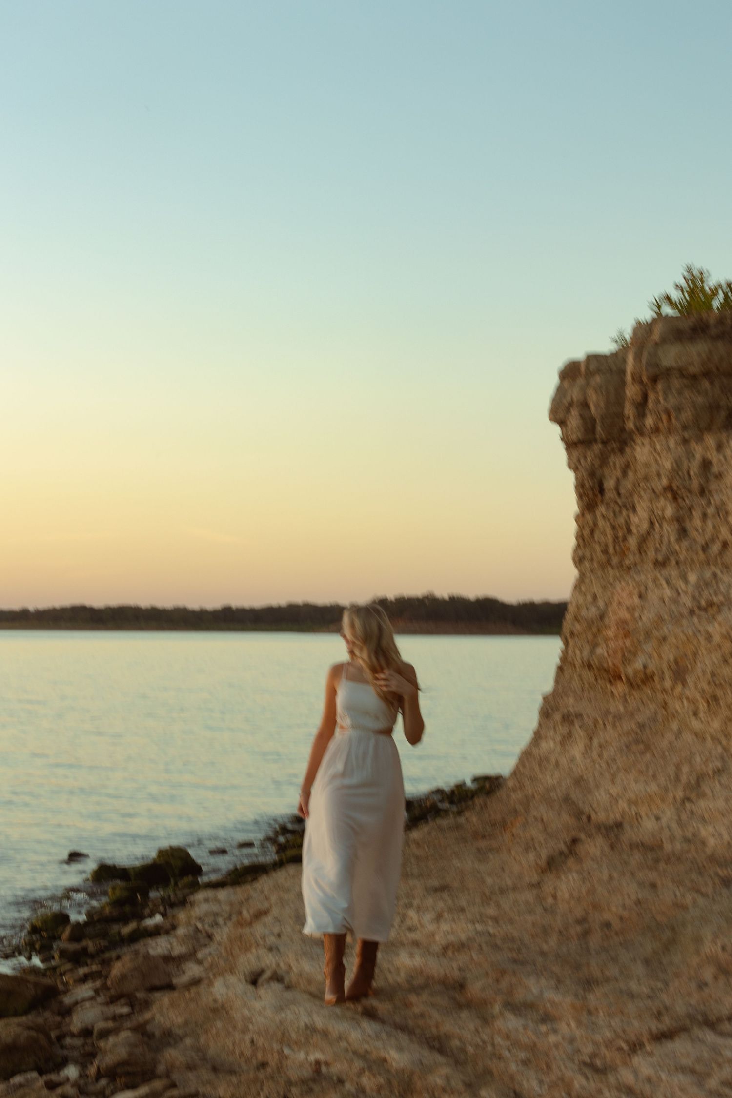 Woman in white dress and boots standing by rocky cliff at golden hour sunset near calm water.