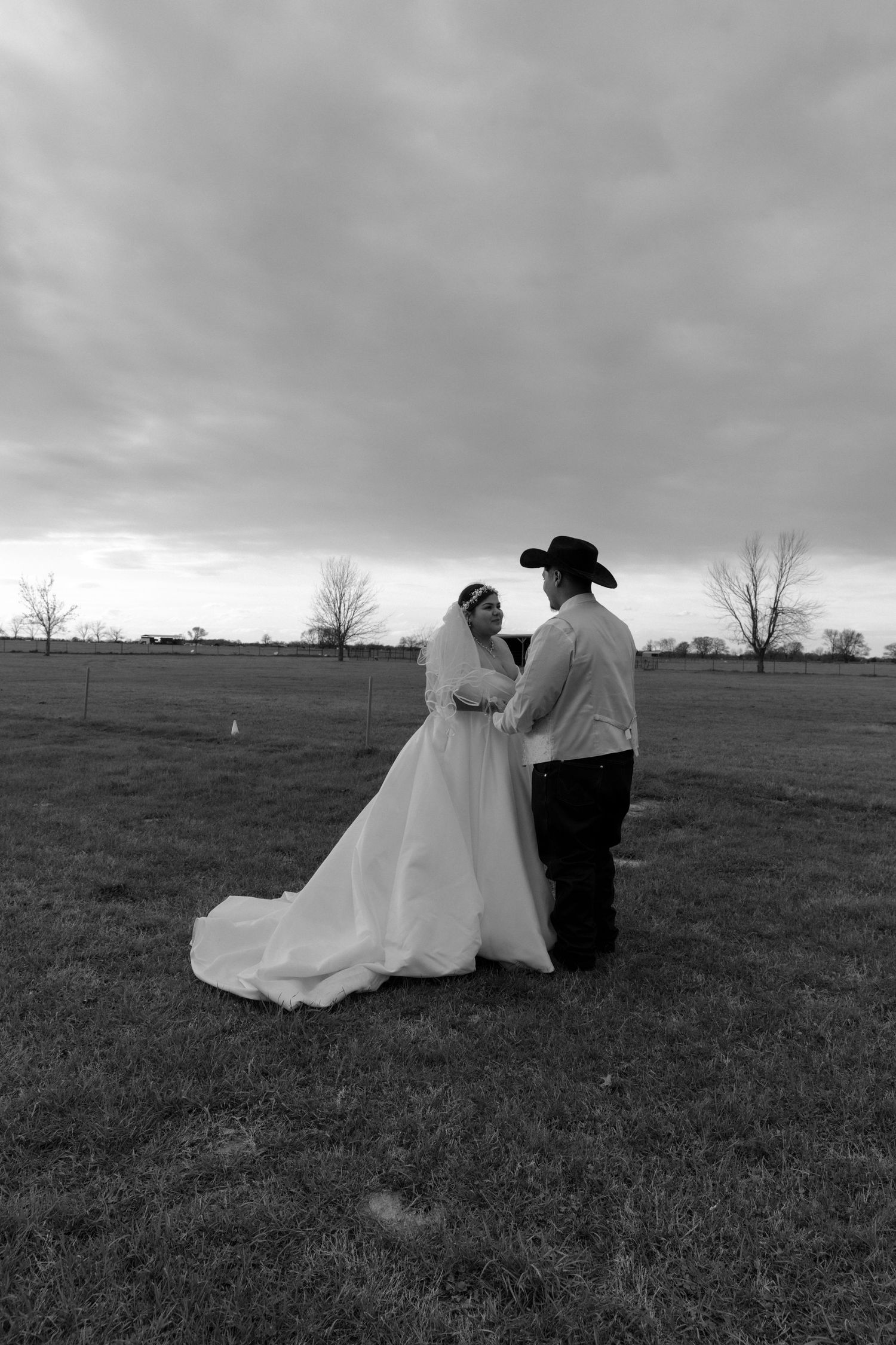 Black and white photo of bride and groom holding hands in an open field under dramatic cloudy sky.