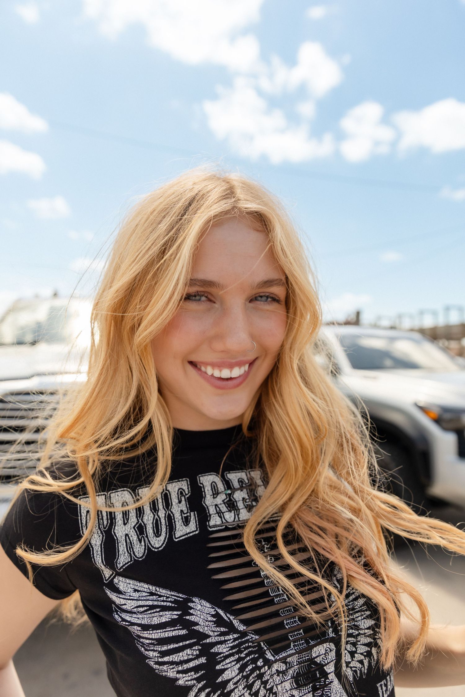 Smiling blonde woman in True Religion graphic tee poses outdoors under blue sky near vehicles.